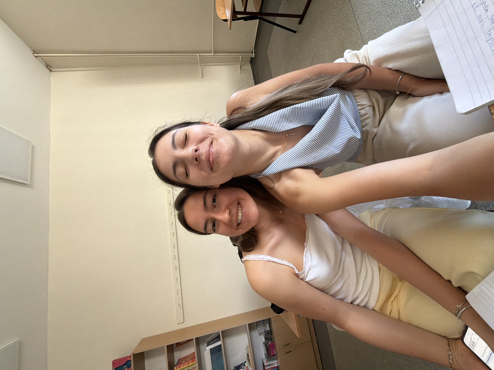 The media file is a portrait-style, slightly overexposed, vertical photograph taken indoors. Two young women are the subjects, positioned close together and leaning into each other, creating a playful and intimate pose. The woman in the foreground, on the right, is looking directly at the camera with a wide, genuine smile, her eyes crinkled at the corners. She is wearing a white tank top with lace trim and a light yellow skirt or shorts. She has a delicate necklace around her neck and several bracelets on her left wrist, one of which is a silver heart charm. Her right arm is wrapped around the other woman.
The woman behind her, on the left, is also smiling, with her eyes closed and her head tilted back slightly. She is wearing a blue and white striped off-the-shoulder top and a white outfit underneath. Her hair is long and brown, flowing down her back. Her left arm is resting on the other woman's shoulder.
The background appears to be a classroom or study area. To the left, there is a plain, light-colored wall with a white rectangular object mounted on it. In the center and to the right, there are bookshelves filled with books, and some papers are visible. Part of a wooden chair is also visible in the upper left corner, and a notebook with handwritten text is in the upper right. The overall impression is one of friendship and happiness, likely during a study break or a casual moment. The lighting suggests it is daytime, and the indoor setting hints at a school or university environment.
Visible text includes handwritten notes on a piece of paper in the upper right corner, but the content is not legible enough to decipher. There is also some text on the spine of books on the shelf, but it is too small to read. The metallic ruler-like markings on the wall to the left of the woman in the foreground are also a notable detail.