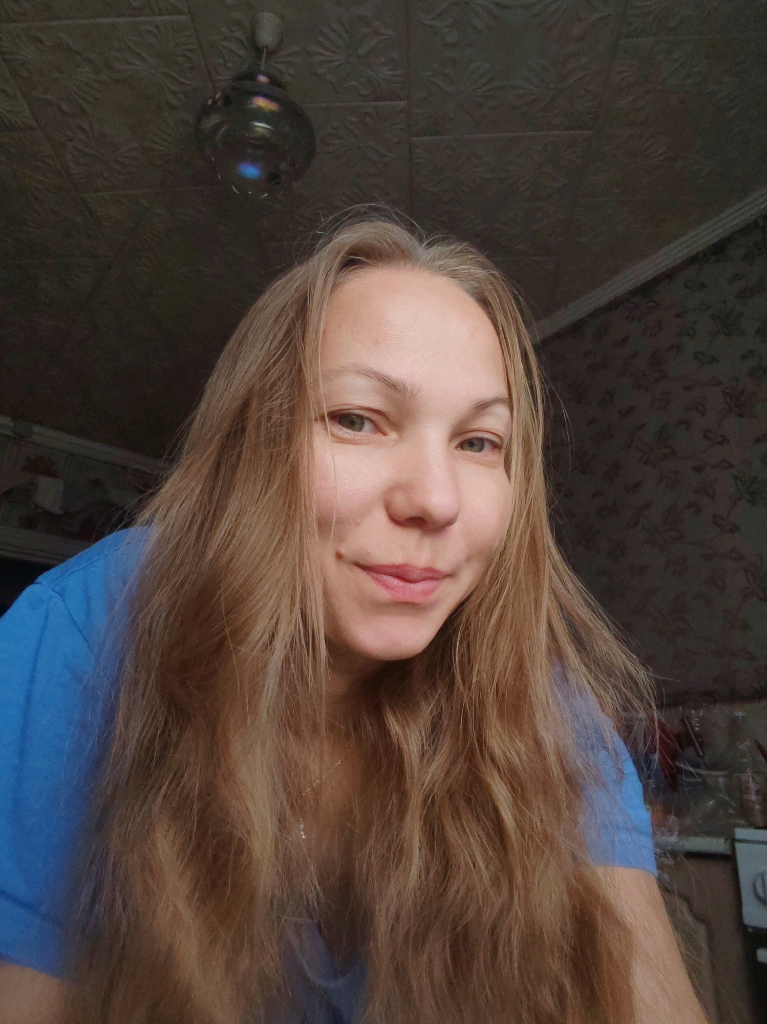A medium shot, taken from a slightly low angle, shows a woman with long, wavy blonde hair and light skin. She is wearing a bright blue t-shirt. Her gaze is directed towards the camera with a slight smile. The background consists of a textured, dark ceiling with a light fixture and a patterned wallpaper. The overall lighting suggests an indoor setting, possibly a domestic environment. The woman's face and hair dominate the foreground, with the background out of focus.