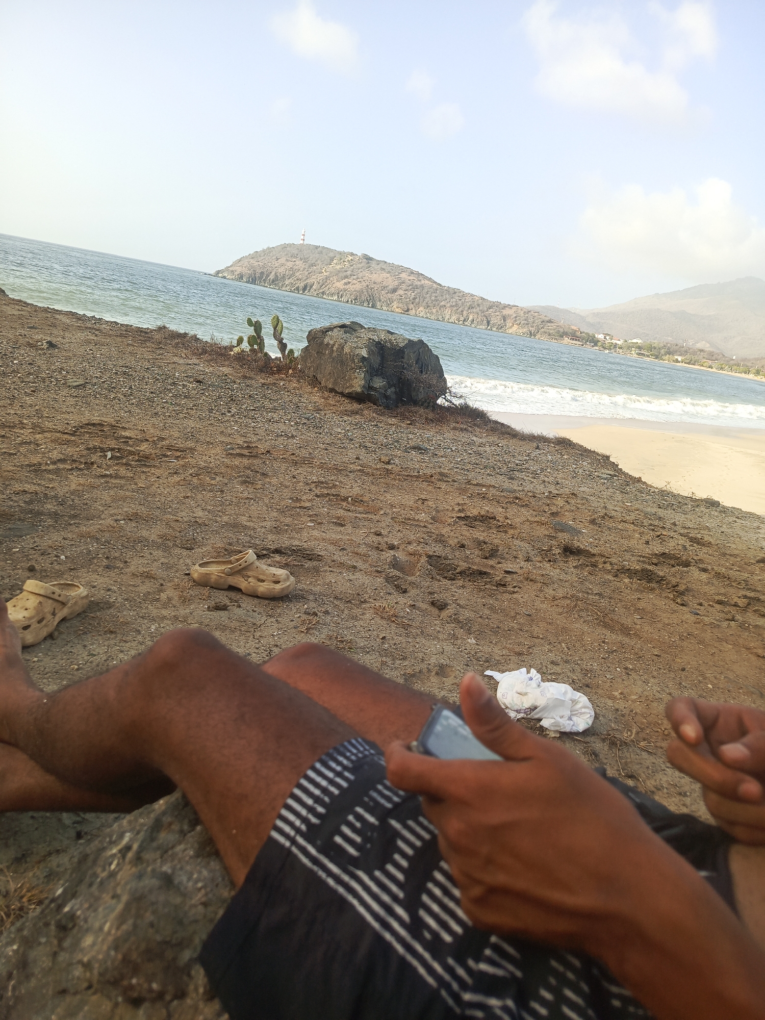 This media file captures a serene coastal scene in Pedro González, Venezuela, from a low, first-person perspective, suggesting the viewer is a person relaxing on the ground. The foreground features the legs and hands of an individual, wearing black shorts with a white patterned design. The person is holding and looking at a smartphone. A pair of worn, light-colored Crocs shoes are placed on the dry, pebbly ground to the left. A crumpled white object, possibly a piece of paper or tissue, lies near the person's hands.
The setting is an arid coastline with dry, earthy ground in the immediate foreground, leading down to the sea. A small cluster of green cacti grows near a large, dark rock closer to the water. The sea itself is calm, with gentle waves breaking on a sandy beach that stretches into the distance. Across the water, a prominent, dry, hilly landmass rises, topped with a distinct red and white striped structure, likely a lighthouse or a communication tower. In the background, further hills and mountains are visible under a bright sky with scattered clouds.
The image conveys a sense of relaxation and leisure on a clear, warm day, likely late morning or early afternoon given the soft lighting. No visible text is discernible on the phone screen or elsewhere in the image.