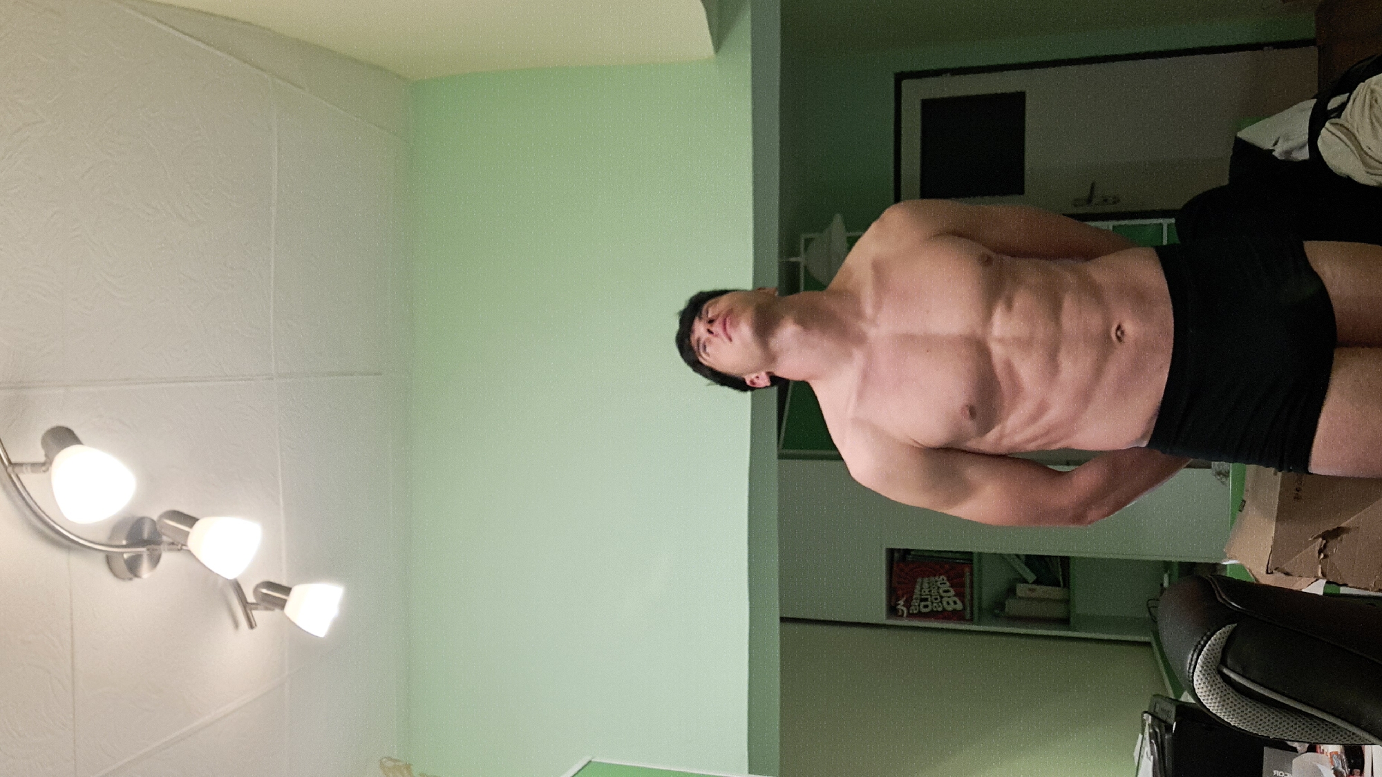 A shirtless man wearing black shorts is standing indoors. The room has white and green walls. There is a multi-bulb lighting fixture on the white wall. A doorway and shelving are visible in the background. A chair and boxes are also present. The location is indoors and there is no information to confirm it is in Burgas, Bulgaria.