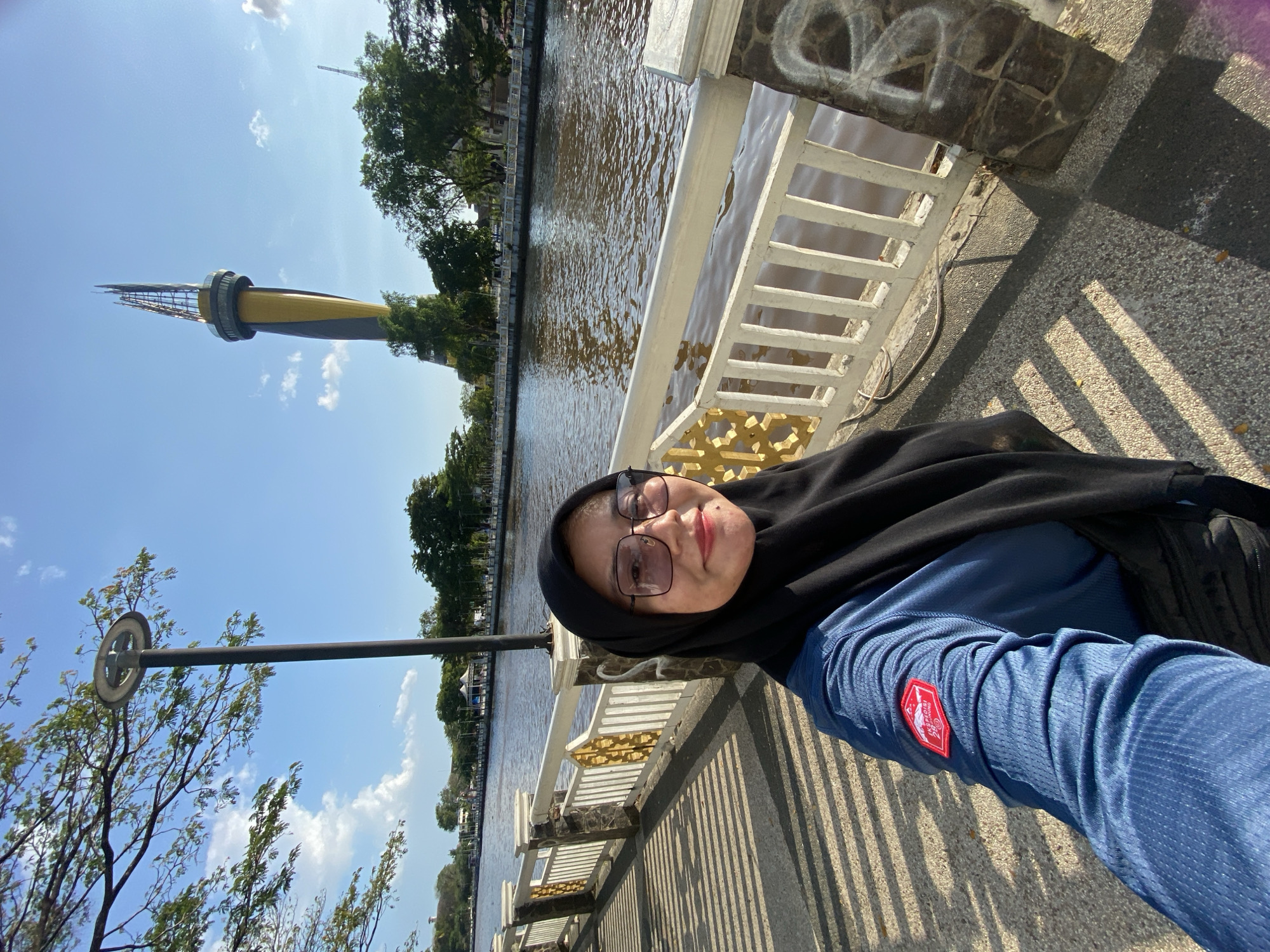 A woman wearing a black hijab and a blue long-sleeved shirt takes a selfie on a paved walkway next to a river. She is smiling and wearing glasses. A white railing with decorative gold elements runs along the river. In the background, there is a tall, distinctive yellow and grey tower, trees, and buildings. The sky is blue with some clouds. The walkway has dappled sunlight creating striped shadows. The scene appears to be located in Banjarmasin, Indonesia, likely near the Siring Pierre Tendean, a popular riverside promenade.