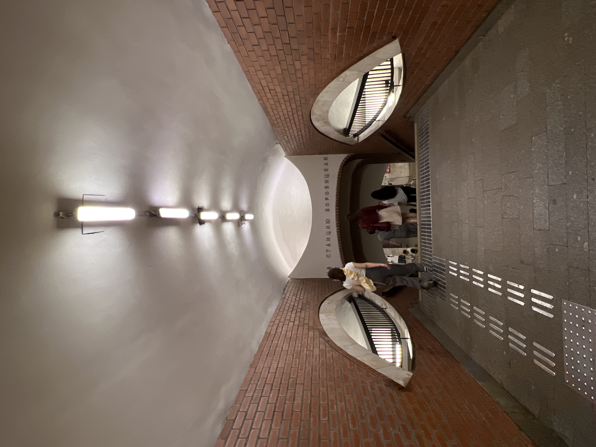 The image depicts an interior view of a subway station in Moscow, Russia. The perspective is from a walkway leading down a set of stairs, with people ascending. The architecture features curved white ceilings with a series of fluorescent lights. The walls are covered in red brick, and there are two distinctive arched openings on either side, resembling eyes, each containing a black metal railing and illuminated from behind. Above the entrance to the stairs, in Cyrillic text, is the station name "СТАНЦИЯ БОРОВИЦКАЯ" (Station Borovitskaya). The walkway is paved with stone tiles, and tactile paving is visible on the ground, indicating it is a public space designed for accessibility. Three individuals are partially visible ascending the stairs: a woman in a white top and dark pants carrying a yellow bag on her left shoulder, followed by another woman in a red top and white skirt, and a third person in dark clothing. The lighting is artificial, casting shadows and highlighting the textures of the brick and ceiling.