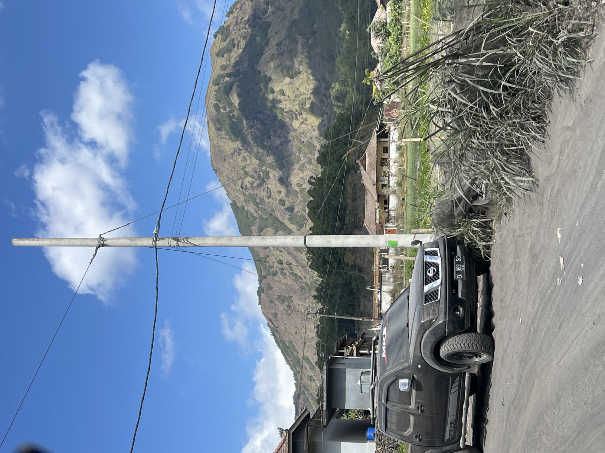 The image shows a dark-colored Nissan Navara pickup truck parked on a dusty, unpaved road. The truck has a license plate that reads "DK 999 SC". Behind the truck, there are several houses with traditional Balinese architecture and thatched roofs. In the background, a large, verdant mountain dominates the landscape, with some trees visible on its slopes. The sky is blue with scattered white clouds. The scene appears to be a rural area, possibly in Kintamani, Indonesia, given the architectural style and the presence of the mountain. The overall impression is one of a serene, natural environment with a touch of human habitation.