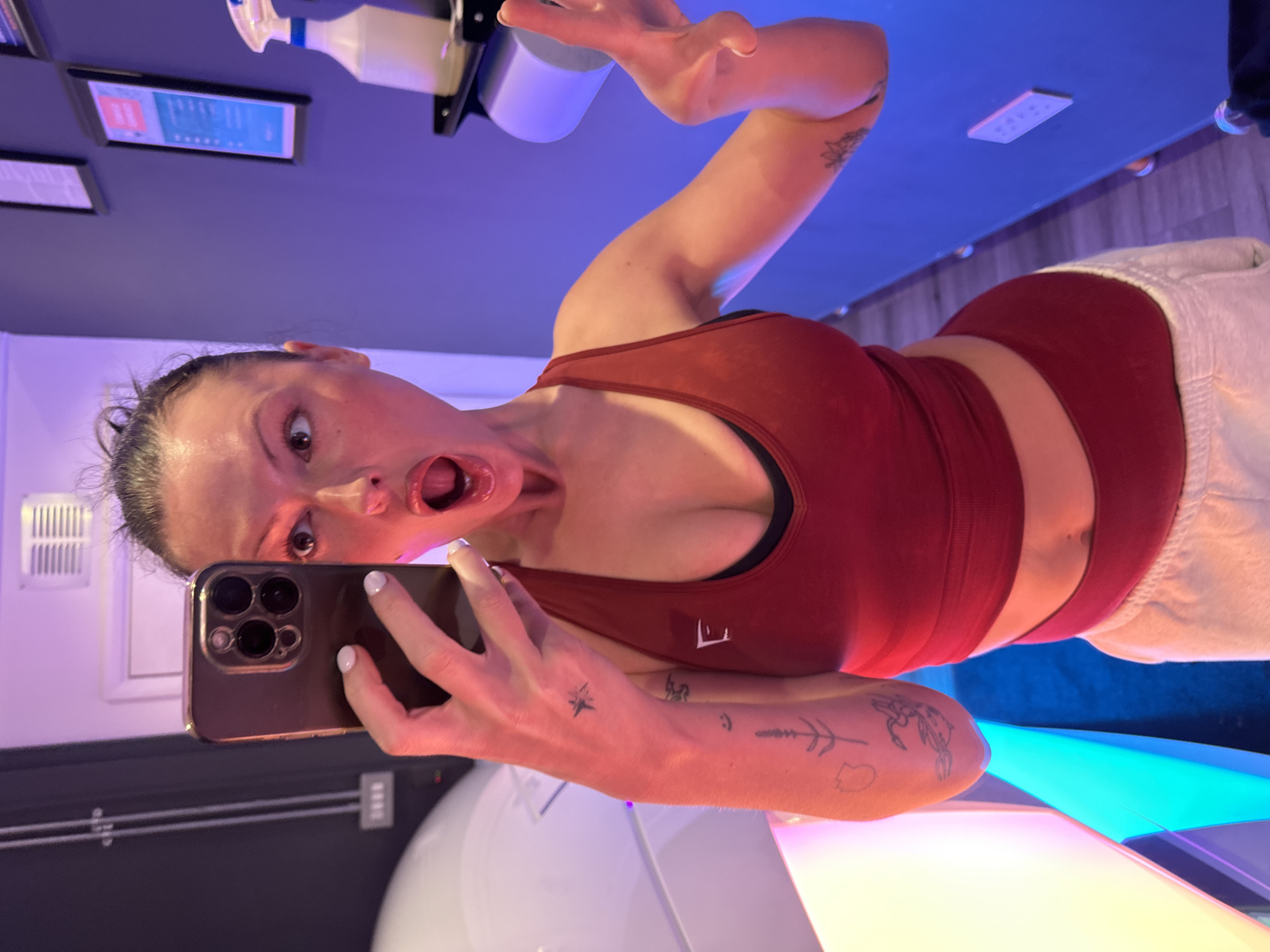 A female subject is depicted taking a mirror selfie within what appears to be a commercial establishment, specifically a tanning or wellness center, located in Tunbridge Wells, United Kingdom. She wears a red cropped sports bra, revealing her midriff, and matching high-waisted red athletic shorts, partially covered by light-colored sweatpants. Her hair is pulled back, and her face exhibits an exaggerated expression with a wide-open mouth and wide eyes. She holds a dark-colored iPhone with a triple-lens camera array in her right hand, capturing her reflection. Her left hand is raised, forming a two-finger peace sign. Multiple small, dark-ink tattoos are visible on her forearms, including a starburst, a botanical stem, and abstract shapes. Her fingernails are painted white.
The environment features a prominent, curved white apparatus on the left, possibly a tanning bed or a floatation tank, emitting pink, blue, and yellow light. The walls are painted a uniform purplish-blue. On the right, a dark shelf holds a white spray bottle with a blue nozzle and a roll of white paper, likely a dispenser. Several framed notices or informational signs are mounted on the blue wall. The overall lighting is low, primarily provided by the colored accent lights from the apparatus and a ceiling light visible in the top center.