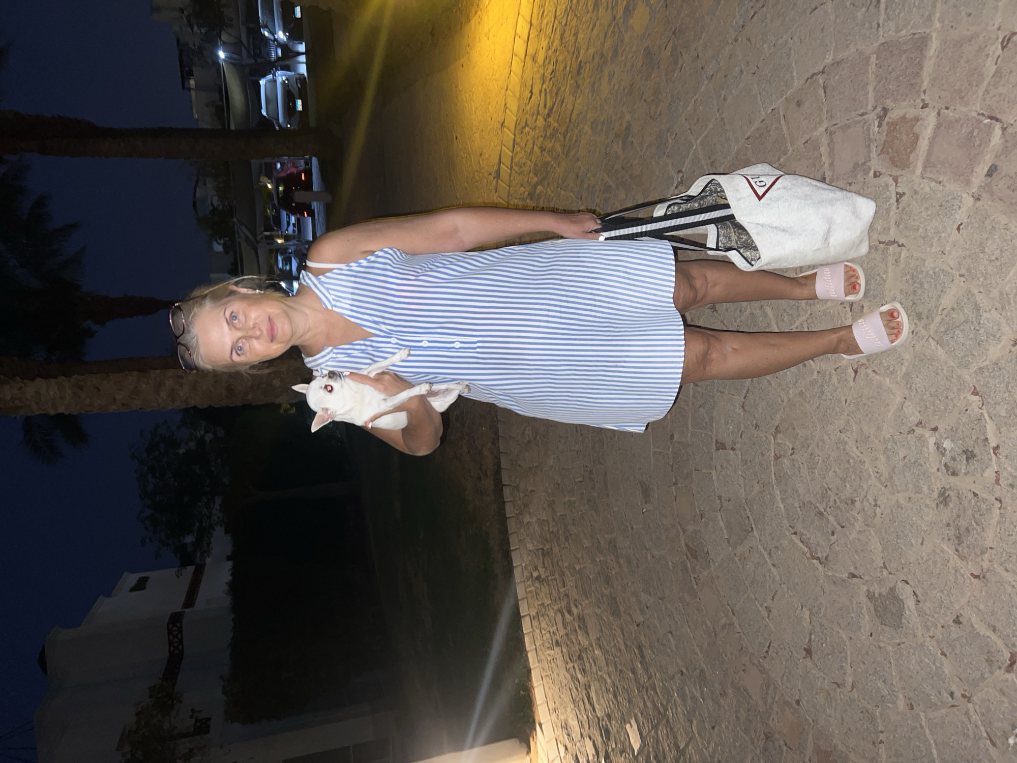 An adult Caucasian woman with blonde hair and blue eyes stands outdoors at night, holding a small white chihuahua. She is wearing a blue and white striped sundress, pink Adidas slides, and glasses perched on her head. She is also carrying a white tote bag with red lettering. The ground is paved with cobblestones, and palm trees and a building are visible in the background. Cars are parked in the distance, illuminated by streetlights. This scene appears to be in Sharm El Sheikh, Egypt, given the presence of palm trees and the architectural style of the building, consistent with resort areas in that region.