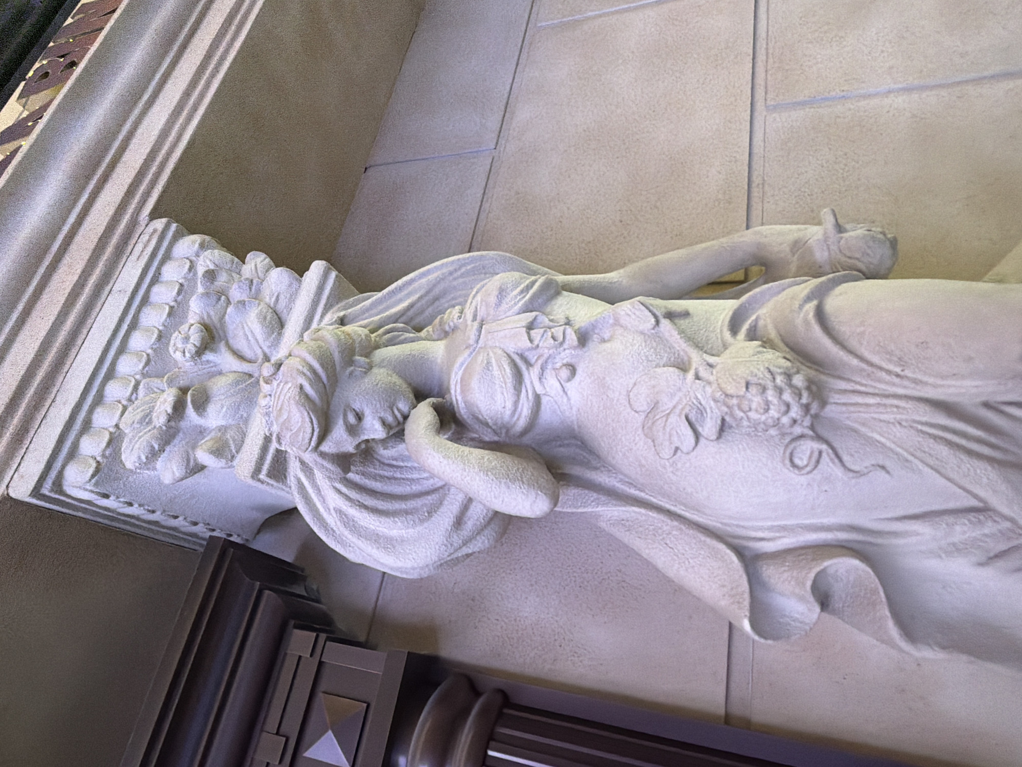 The image shows a stone sculpture of a woman holding a bunch of grapes. The sculpture is a decorative element, likely part of a building's facade or interior design. The woman is depicted with flowing hair and draped clothing, characteristic of classical or neoclassical styles. She has a pensive expression and her hand is near her face. Behind and to the left of the sculpture, a dark, paneled architectural feature is visible, possibly a column or a decorative recess. The background wall is textured and appears to be made of stone or stucco. The lighting suggests an indoor setting or an overcast day. There are no people visible in the image, only the sculpted figure. The scene does not provide any information that would allow for its location to be identified in Ulaanbaatar, Mongolia.