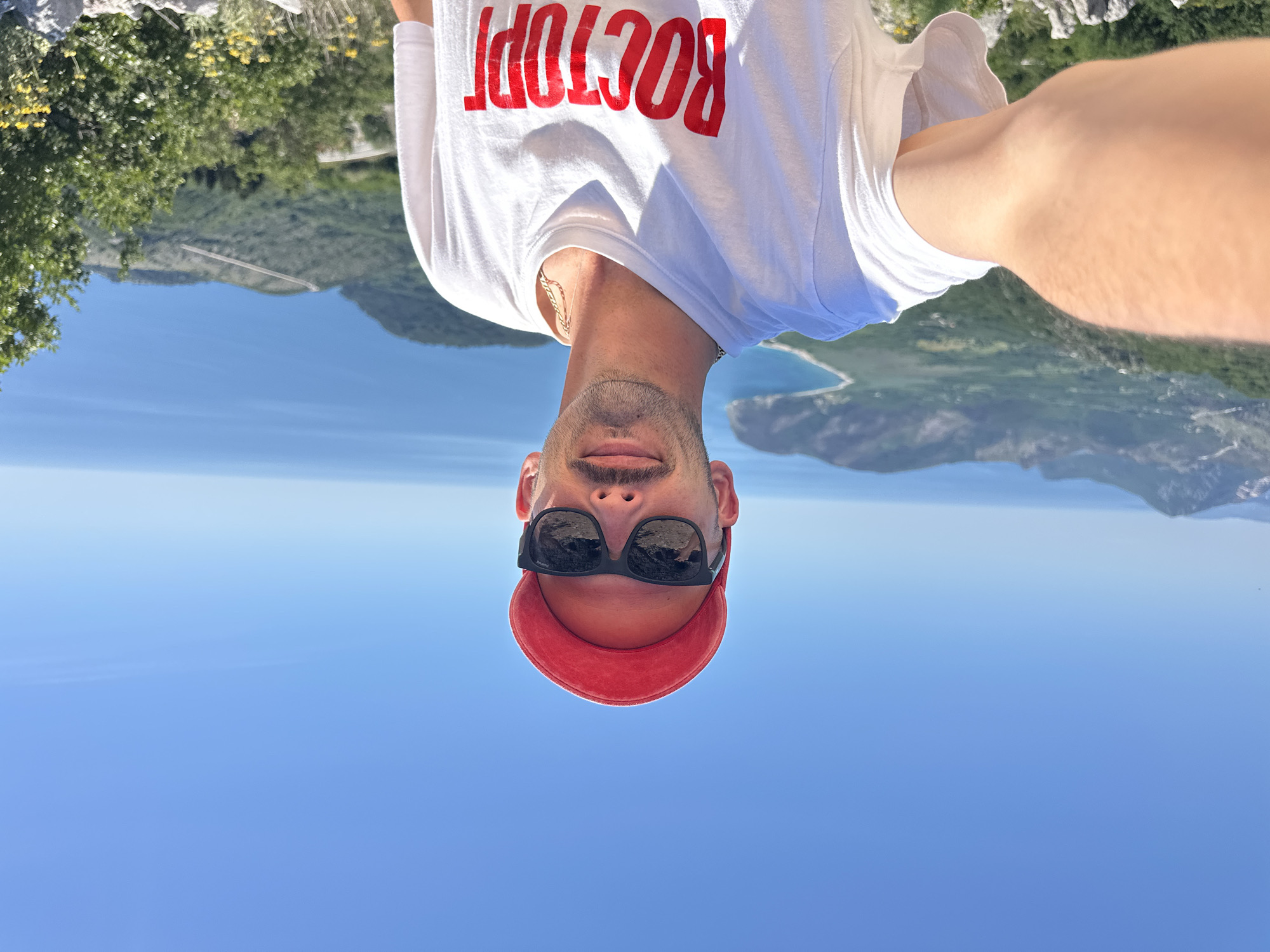 A selfie photograph shows a man wearing a white T-shirt with the word "BOCTOPL" in red letters, a red cap, and sunglasses. The background includes a blue sky, a body of water, and green vegetation. The location is Novoselje, Montenegro.