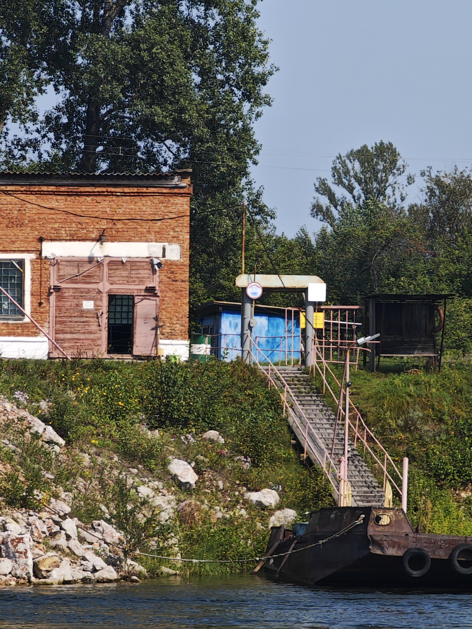 The image depicts a waterfront scene in the village of Esaulovo, Russia, on a sunny day with a clear blue sky. In the foreground, a rusty, flat-bottomed barge is moored in the water, with several tires attached to its side for protection. A rope extends from the barge to the shore.
To the left of the barge, a steep, rocky embankment rises, covered in green vegetation and scattered wildflowers. Atop the embankment, a weathered, multi-story brick building with a corrugated metal roof is visible. This building has large, wooden double doors, one of which is ajar, revealing a darker interior with a window. A single window with a metal grate is also visible on the side of the building. A pink pipe runs along the exterior wall of the building.
Adjacent to the building, a set of stairs ascends the embankment, leading to a small platform with a light blue structure and a darker wooden shed behind it. The stairs have pink railings. A sign with a red border and a white circle containing a red wave symbol is mounted on a pole near the top of the stairs. The surrounding area is lush with trees and foliage. The overall impression is one of an industrial or utility area situated by a body of water, likely a river or lake.