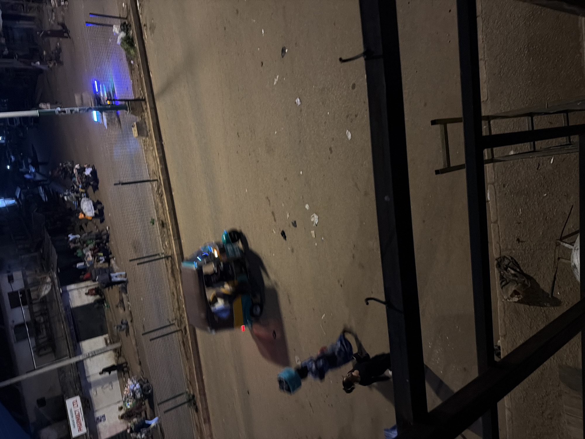 The image depicts a street scene at night in Bukuru, Nigeria. In the foreground, a metal railing separates the viewer from the street below. A woman is seen walking with a blue basket on her head, wearing a blue dress. To her left, another person in a dark outfit is looking down at their phone or an object in their hand.
In the middle ground, a three-wheeled auto-rickshaw, commonly known as a "Keke Napep," is moving across the street. It has a yellow and blue color scheme and appears to have lights illuminated, casting a blue glow. The Keke Napep is carrying passengers, though their identities are not clear due to the low light and motion blur.
The background shows a building with signs, one of which reads "PHARMACY LTD." Several people are standing or walking near this building, and other vehicles, possibly motorcycles, are parked. The street itself is paved and has some scattered debris. The overall atmosphere is that of a busy urban environment at night.