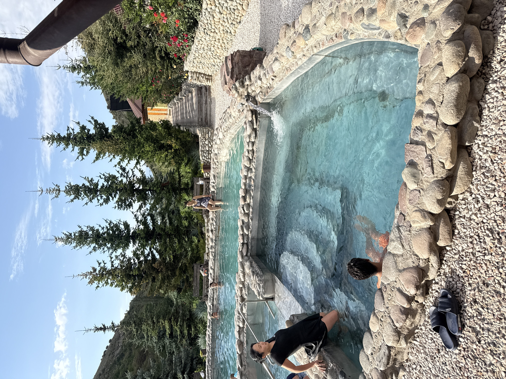The image depicts an outdoor hot spring facility located in Ak-Suu, Kyrgyzstan. Several people are visible in and around the turquoise water pools, which are bordered by natural stone. In the foreground, a woman in a black dress is partially submerged in a pool, holding onto the edge. To her right, a person with dark hair is relaxing in the water, facing away from the camera. In the background, other individuals are also enjoying the hot springs. The surrounding environment includes lush green trees, a hillside, and a clear blue sky with scattered clouds. A stone pathway and a building with a reddish roof are visible further in the distance. The ground around the pools is covered in small pebbles.
