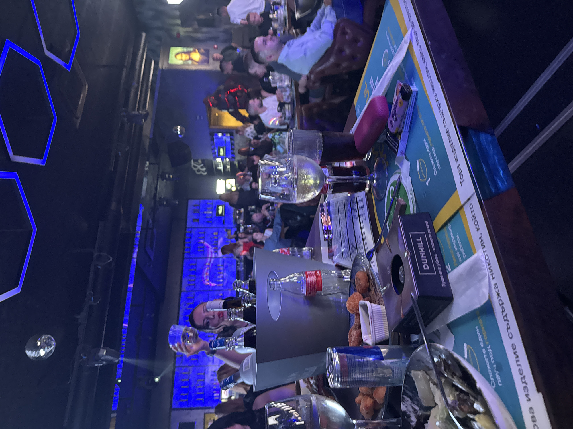The image displays an indoor nightlife venue in Sofia, Bulgaria, evidenced by Bulgarian health warnings printed on table coasters. The scene is dimly lit with prominent blue LED lighting on the ceiling, where hexagonal panels emit light, and along a lengthy bar counter in the background. A disco ball is suspended from the dark ceiling.
In the foreground, a wooden table holds a grey ice bucket containing several dark glass bottles and a clear plastic Coca-Cola bottle. Adjacent are a clear wine glass filled with a light-colored liquid, a tall glass with a dark beverage, and a blue Red Bull can. A plate contains fried food items and a small white dish with dipping sauce. Other objects on the table include a black Dunhill box, a pink item, a patterned cigarette lighter, and white napkins. Paper coasters beneath some items display Bulgarian text warnings concerning nicotine and smoking.
A woman with dark hair, wearing a white top and red lipstick, is positioned in the mid-foreground, holding a clear glass above her head while looking towards the viewer. In the midground and background, approximately 20 to 30 other individuals are visible, primarily seated at tables or standing near the bar. A man in a light blue shirt is clearly discernible in the midground right, seated in a brown leather chair. Further back, a figure in a dark outfit with red illuminated accents is present. The background features a well-stocked bar area with blue backlighting and a large illuminated portrait resembling the Mona Lisa on the right wall.