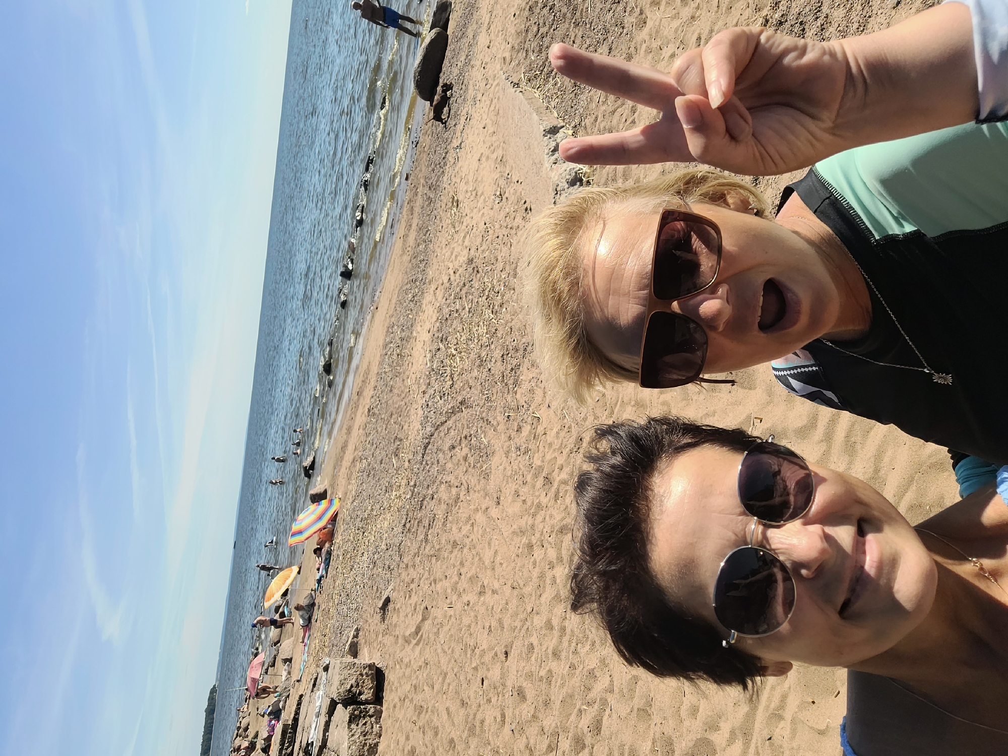The image shows two women on a sandy beach with a body of water in the background. The woman on the left, in the foreground, has dark hair, is wearing sunglasses, and smiling. The woman on the right, also in the foreground, has blonde hair, is wearing sunglasses, and is making a peace sign with her right hand, with her mouth open. She is wearing a black and teal rash guard. In the background, there are several people on the beach, some under umbrellas, and others in the water. There are also rocks along the shoreline. The sky is blue with some clouds. The scene appears to be a sunny day at a beach resort. The location is identified as Repino, Russia.