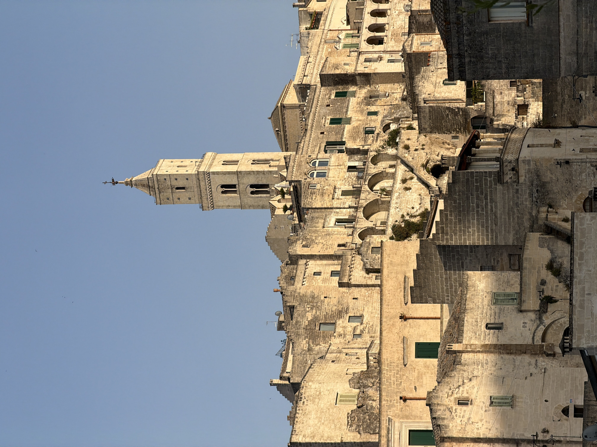The image depicts the Sassi di Matera, an ancient cave dwelling settlement located in Matera, Italy. The scene is dominated by a cluster of stone buildings carved into the hillside, showcasing a complex and layered architecture. A tall, light-colored bell tower with arched windows and a cross-topped spire stands prominently against a clear blue sky, suggesting it is daytime. The buildings are constructed from weathered, light-colored stone, with many featuring arched windows, small openings, and protruding elements. Green shutters are visible on some of the windows. The overall impression is of a historic and rugged urban landscape bathed in natural light, with no people or animals readily apparent.