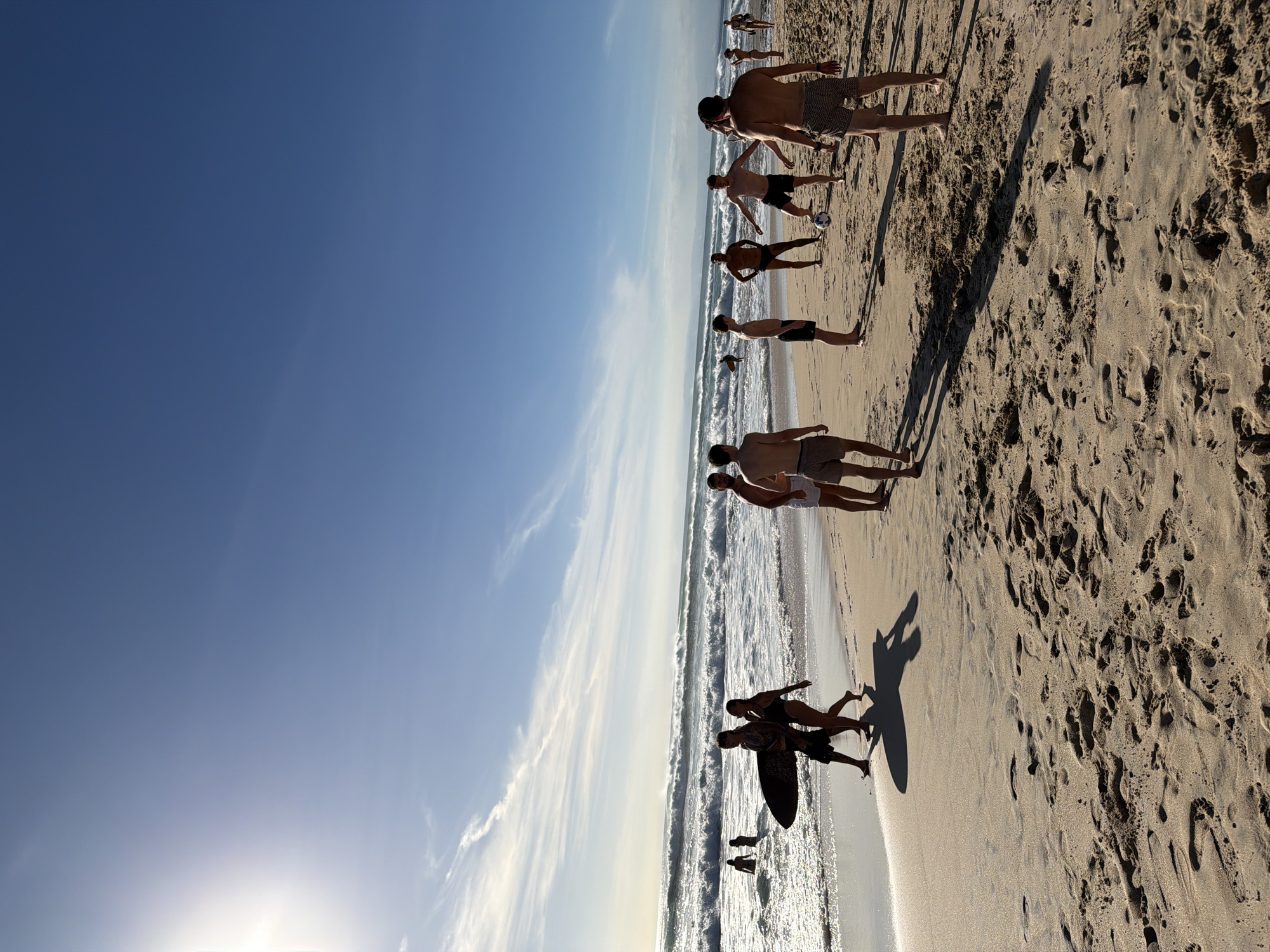 A sunny beach scene in Kuta Selatan, Indonesia, features a group of people on the sand near the water's edge. A cloudless blue sky dominates the upper half of the image. A line of people, mostly shirtless young men, stands parallel to the shoreline. One pair carries a surfboard. The sand is light-colored with numerous footprints. Waves break near the shore.