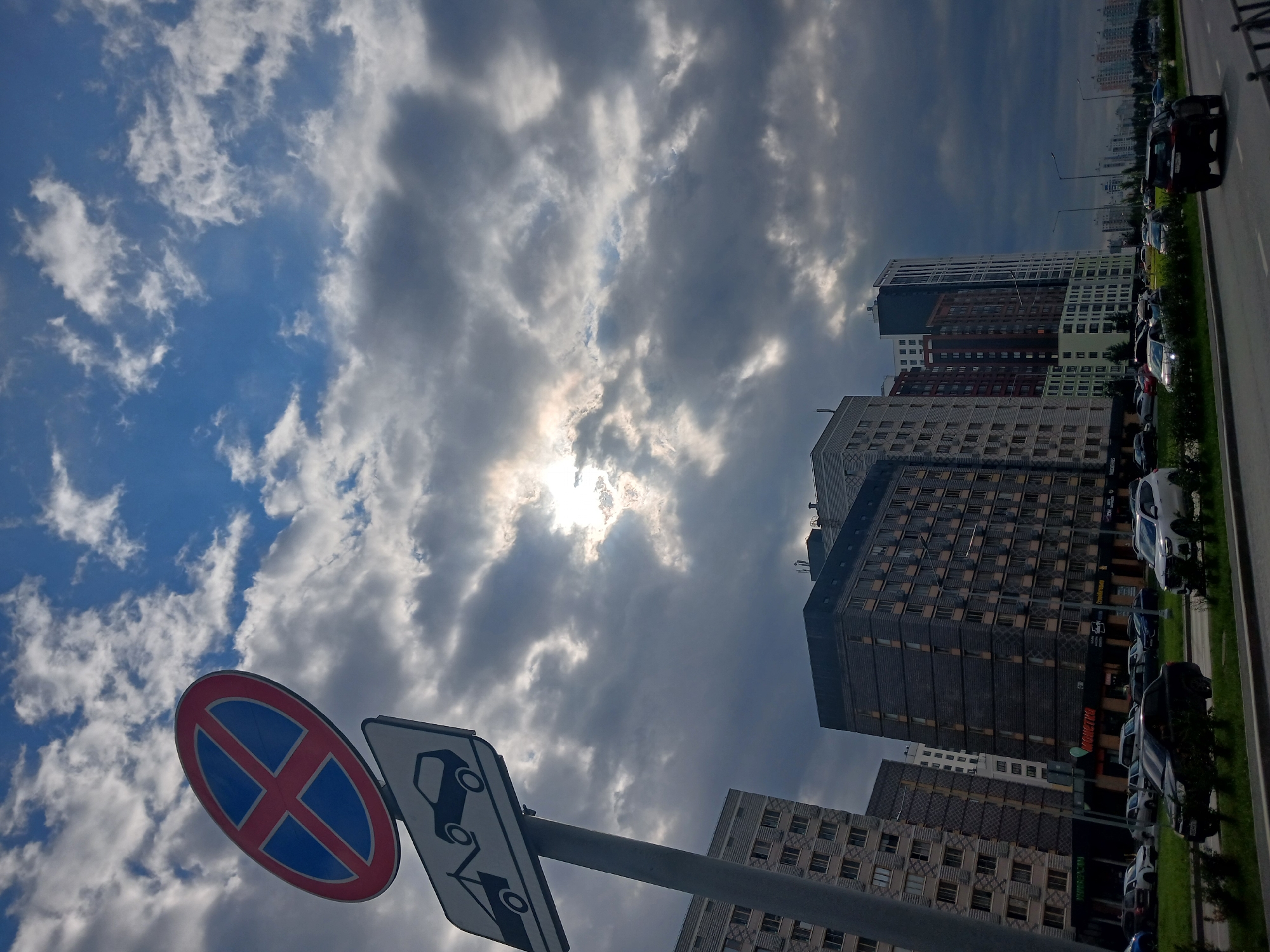 The image shows a street scene in Yekaterinburg, Russia, on a cloudy day with the sun breaking through the clouds. In the foreground, a traffic sign with a red circle and a blue cross indicates no stopping or parking, with a tow truck symbol below it. A gray pole supports the signs. A road runs horizontally across the image with several cars driving on it and parked alongside. In the middle ground, modern apartment buildings of varying heights and architectural styles line the street. One building has a sign for "MONETKA", a Russian supermarket chain. In the background, more buildings stretch into the distance, suggesting a densely populated urban area. The sky is filled with dramatic, dark clouds interspersed with patches of blue sky, and the sun creates a bright, hazy glow. There are no people visible in the image.