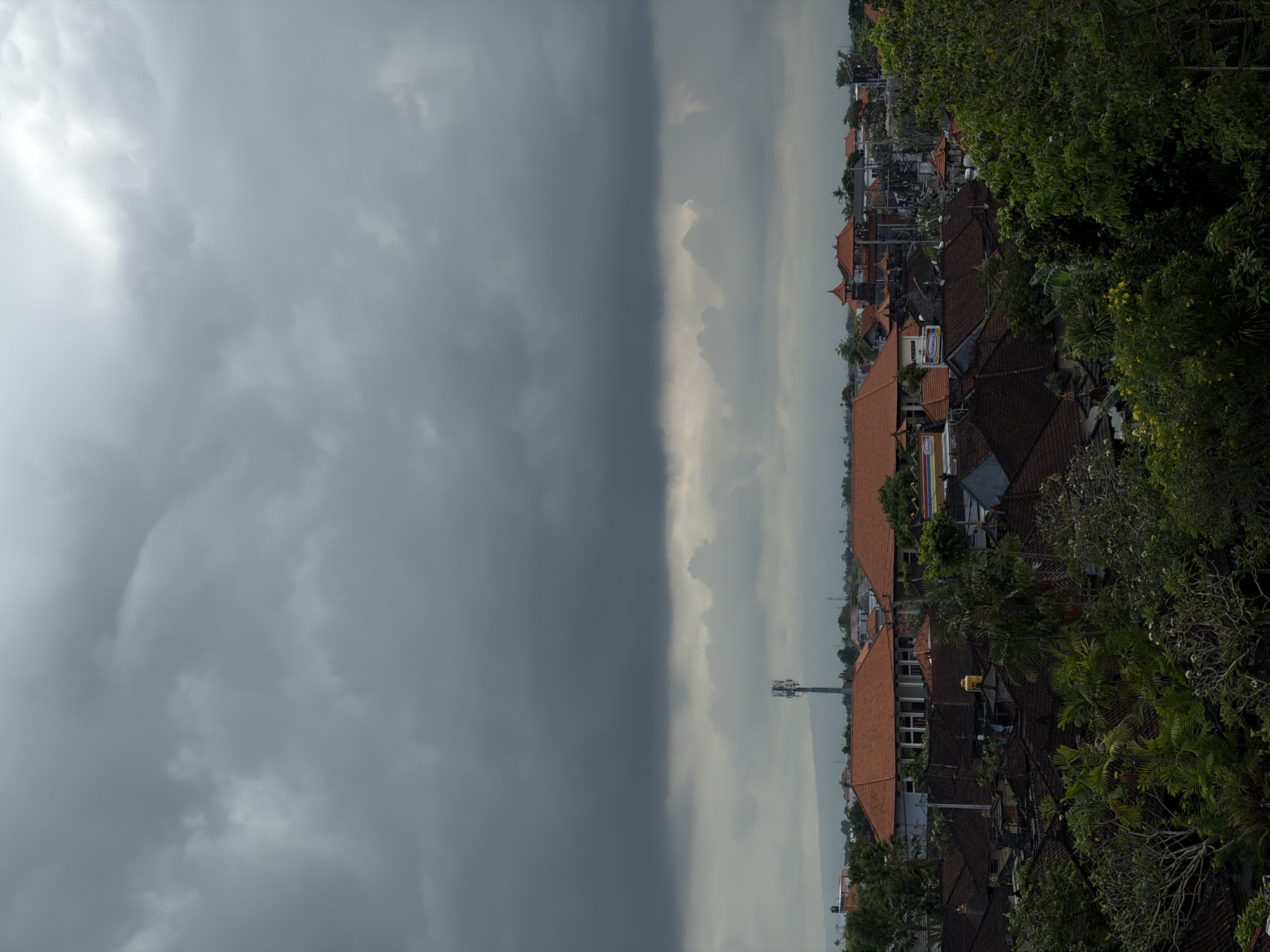 The image captures a distinct weather phenomenon over the city of Mengwi, Indonesia. The sky is bisected: one side is dominated by dark, ominous storm clouds, contrasting sharply with the other side, which is characterized by brighter, overcast conditions. Below the dividing line of weather, the cityscape of Mengwi unfolds. Predominantly, there are houses and buildings with red-tiled roofs, nestled among verdant trees and foliage. A communications tower is visible, rising above the buildings. The scene is still and serene, with no apparent signs of human activity.