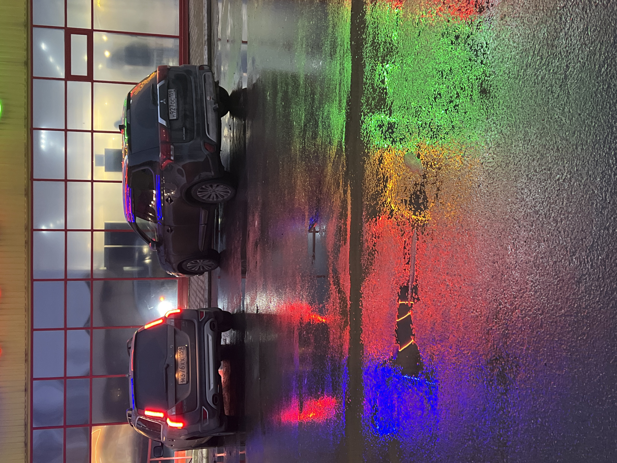 The image shows a wet street scene at night in what appears to be Chaikovsky, Russia, judging by the license plates on the cars. Two grey SUVs are parked near a building with a grid of windows reflecting an internal light. The street surface reflects a spectrum of colors from nearby neon lights or signage: green, yellow, red, and blue. The pavement appears wet from recent rain, enhancing the reflections. Details like the license plate numbers H376XX159 and H927OC159 are visible on the cars.
