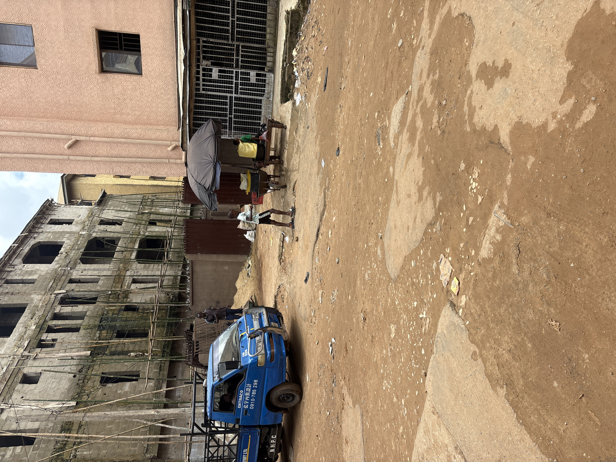 The image depicts a street scene in Onitsha, Nigeria. A blue truck with "ENTRACO" and Chinese characters on its side is parked in the foreground. A person stands next to the truck. In the middle ground, a young boy walks past a market stall covered by a black umbrella. The stall has a wheelbarrow and a table with yellow containers. A person in a green shirt is seated behind the table. Further in the background, a partially constructed building with bamboo scaffolding stands next to a peach-colored building. The ground is a mix of dirt and patches of concrete, with some debris scattered around. The sky is overcast.