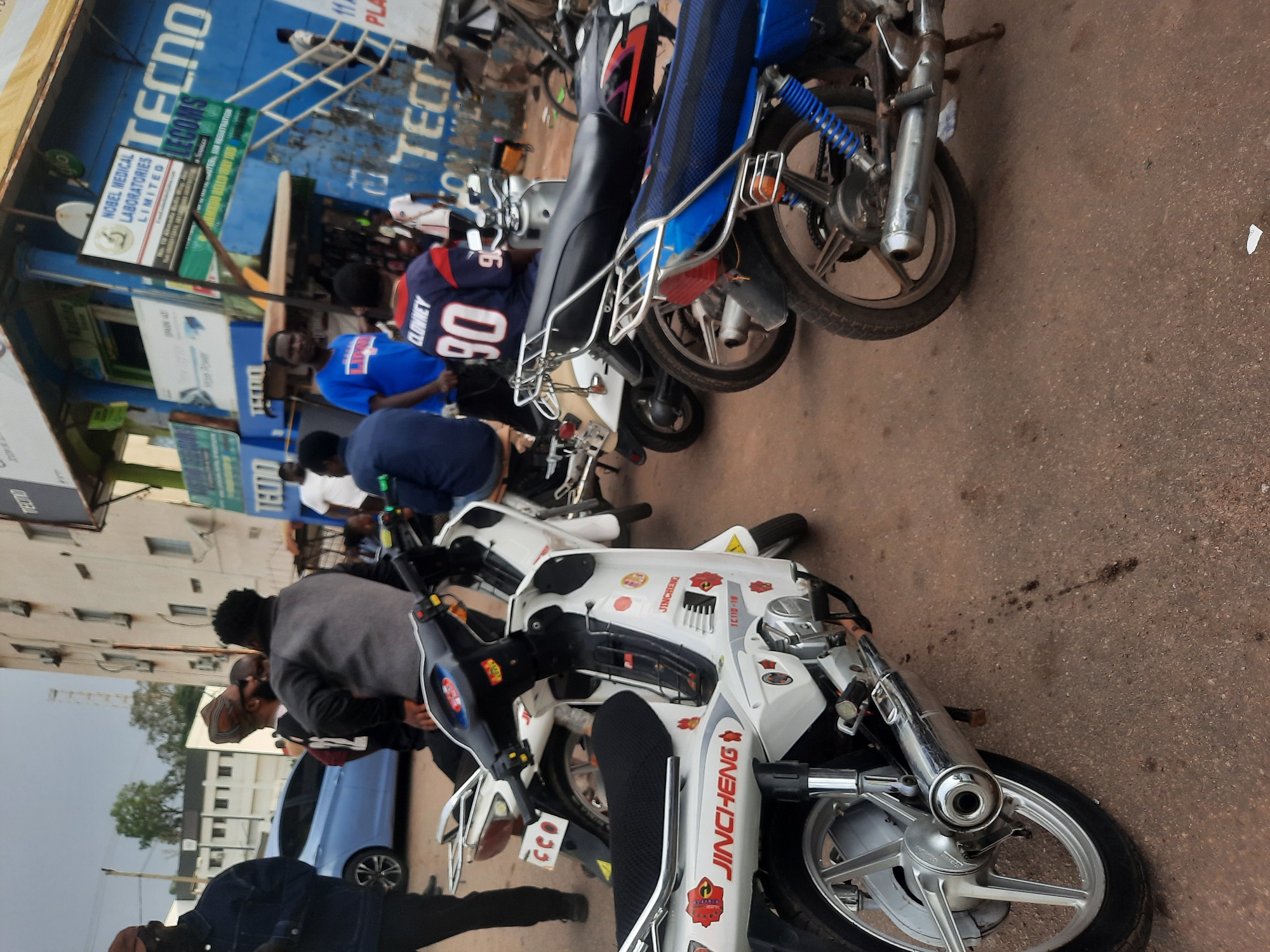 The image shows a street scene in Jos, Nigeria, dominated by parked motorcycles. Several people are gathered near a blue building advertising "TECNO," "LECOMS," and Nobel Medical Laboratories Limited. A man in a blue jersey with the number "90" and the name "CLOWNEY" is visible. Several motorcycles are parked on the right side of the frame. The foreground shows a white "JINCHENG" brand motorcycle. The street surface appears to be paved. A blue car is partially visible.