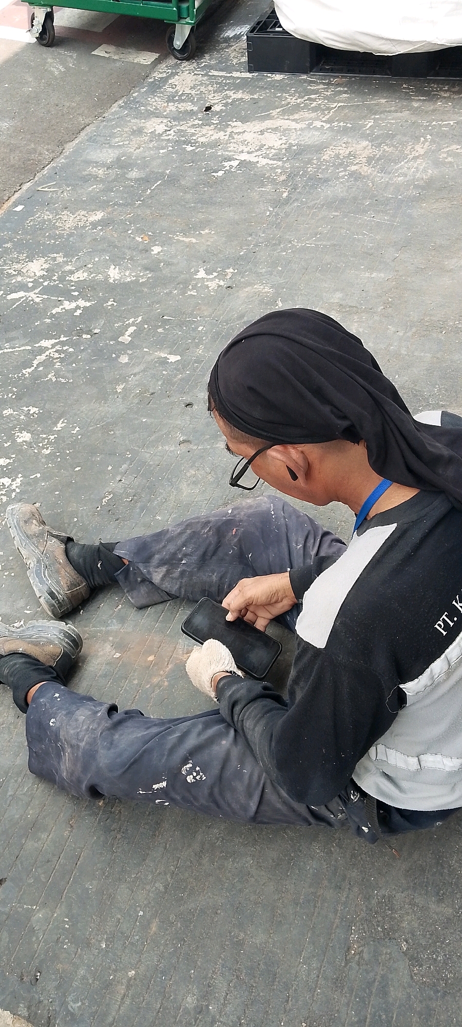 A person wearing dark blue work pants, a black head covering, and a black and white long-sleeved shirt is sitting on a gray concrete floor. The person is holding a black smartphone in their hands, with white gloves on their hands. The person is wearing eyeglasses. The concrete floor appears worn and stained. In the background, a green industrial cart with wheels is visible, as is a white sheet draped over a dark object. The scene is outdoors, possibly in a work or industrial area, likely in Depok, Indonesia, based on the provided context.