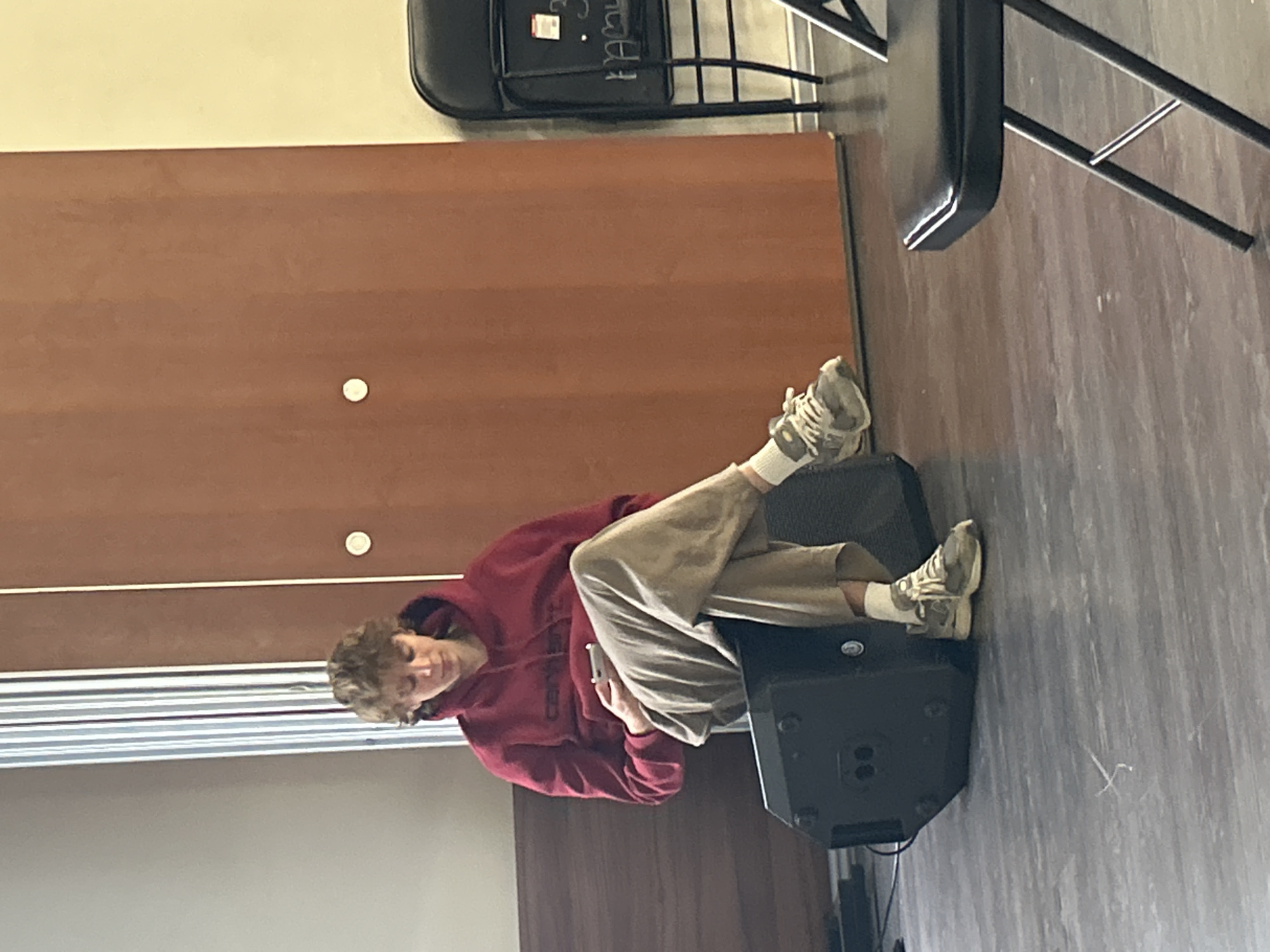 A young person with curly, light brown hair is sitting on a black speaker in a room, looking down at a white smartphone they are holding in their hands. They are wearing a red hooded sweatshirt with the word "carhartt" visible in black lettering on the chest, and loose-fitting beige pants. Their feet are clad in white socks and grey athletic shoes. The room has a light-colored, reflective floor and a large, light brown wooden door in the background. To the right of the door, a black folding chair with a chalkboard attached is visible.