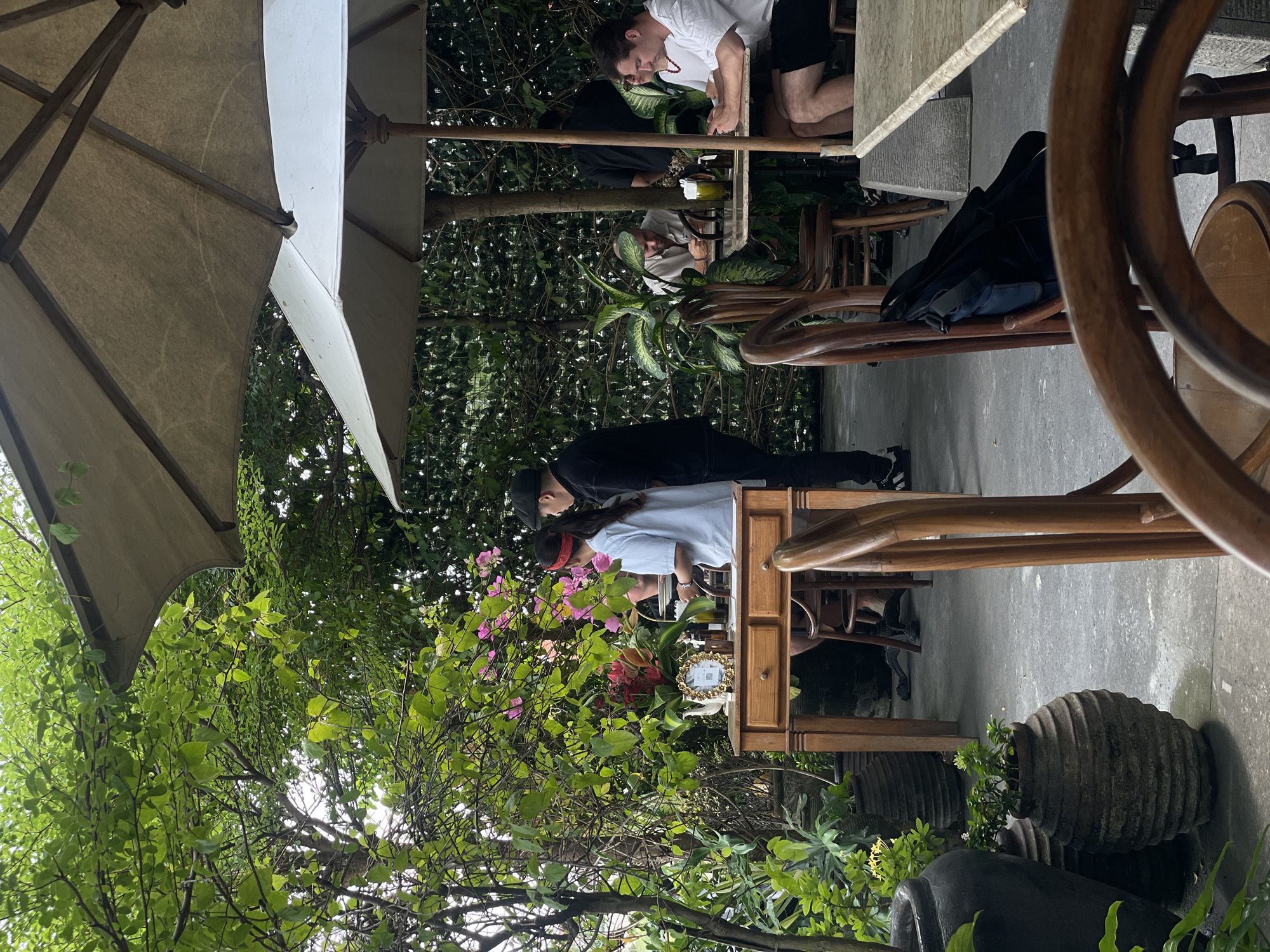 This image captures an outdoor seating area at a cafe in North Kuta, Indonesia. The scene is dominated by lush greenery and large umbrellas providing shade. In the foreground, a wooden table with drawers and a decorative frame holding a picture of a teapot are visible. Two individuals, a woman in a light blue shirt and a man wearing a black cap and shirt, are interacting near the table. Further back, several patrons are seated at tables, enjoying their time. One man, wearing a light-colored shirt, is seated at a table to the left, while two men in white shirts are seated at a table to the right. Large potted plants and wooden chairs are scattered throughout the area, contributing to the relaxed, natural ambiance.
