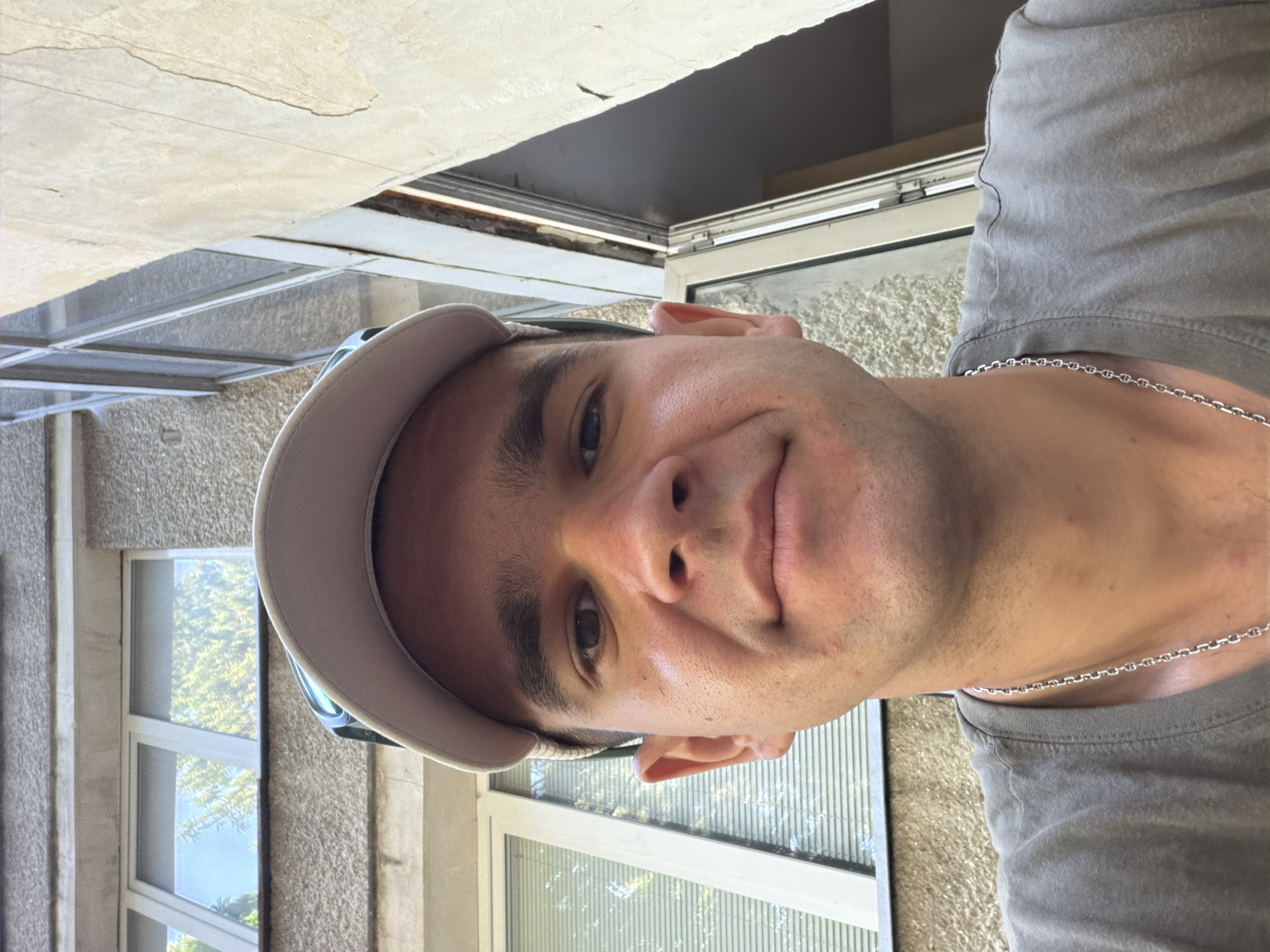 A close-up, low-angle selfie of a man wearing a light-colored baseball cap and a gray t-shirt with a silver chain necklace is taken outdoors. He is looking directly at the camera with a neutral expression. Behind him, a building with textured concrete walls and white-framed windows is visible. Trees and the sky are reflected in the glass of the windows, suggesting it is daytime. The setting appears to be an urban environment, possibly in the city of Veliko Tarnovo, Bulgaria, given the architectural style and the presence of trees and a building facade. The lighting indicates it is a sunny day, with shadows cast on the building. The image is sharp and well-lit, focusing on the man's face and upper torso.