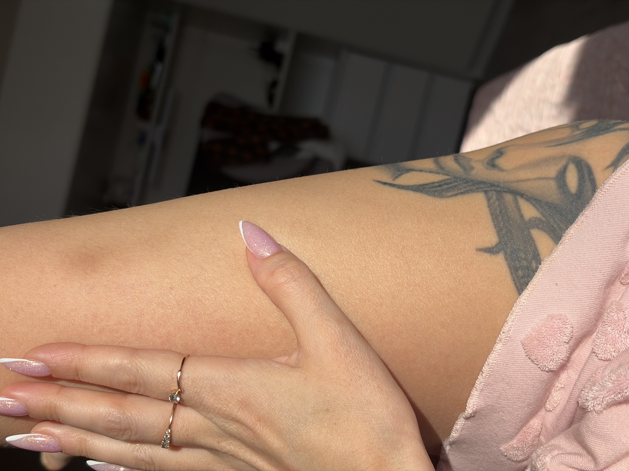 A close-up shot depicts a person's arm and hand. The arm has a tattoo in grayscale. The hand, adorned with two thin, gold-colored rings, has manicured nails with a pink base and a white tip in a French style. The arm rests against a light pink, textured garment. The background is out of focus, but hints of a room with shelves and some light from a window can be discerned. There is no indication of the location in Saint Petersburg, Russia.