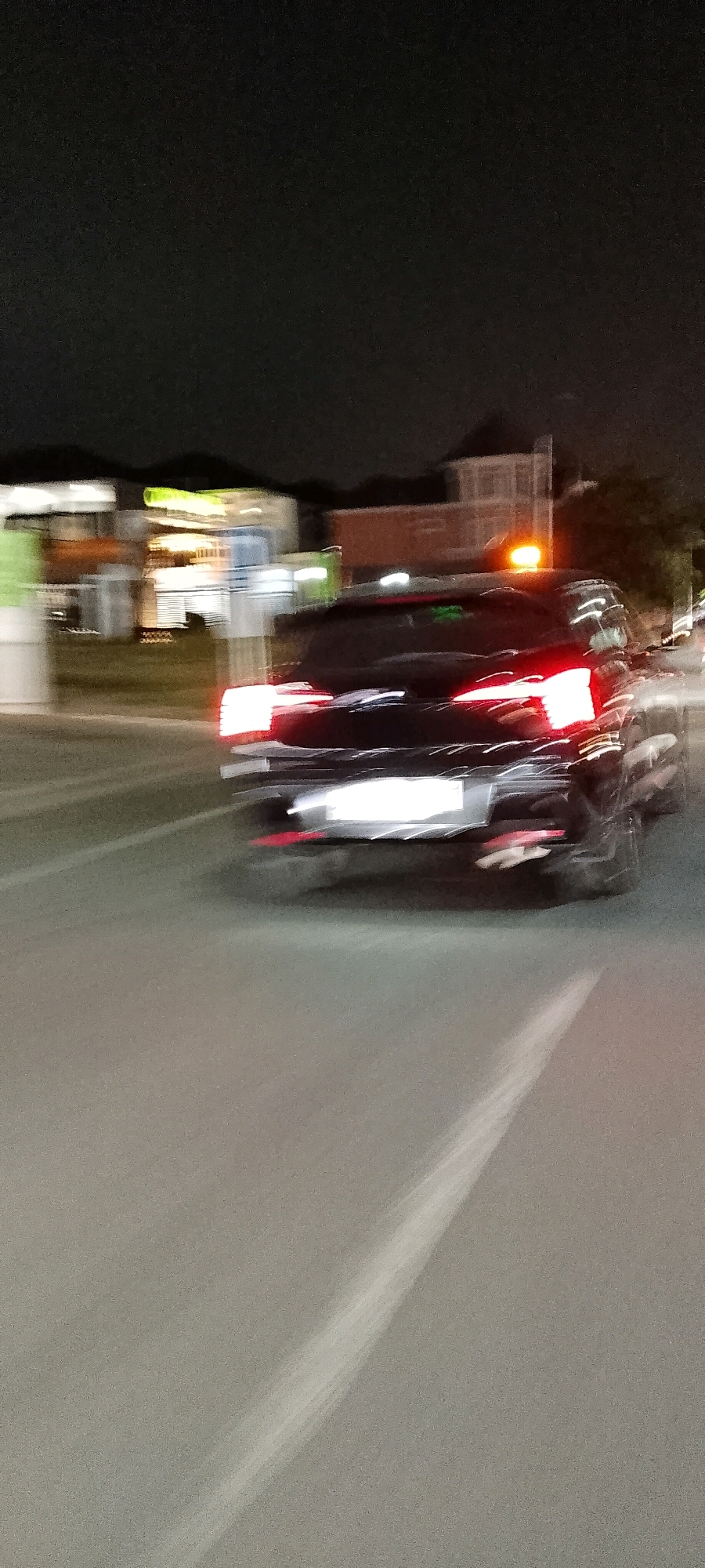 A black vehicle with an orange flashing light on its roof is driving at night on a road in Gunung Putri, Indonesia. The vehicle's taillights are illuminated, and the motion blur suggests it is moving. The surrounding environment appears to be an urban or suburban area with blurred buildings and streetlights visible in the background. The road surface is asphalt, with a visible lane marking.