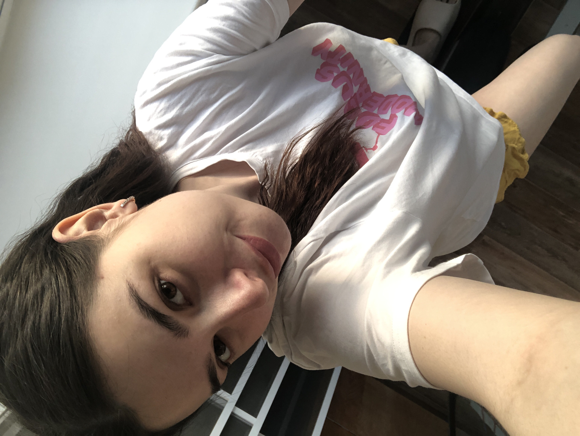 A young woman is posing for a selfie. She is fair-skinned with brown hair and eyes. She is wearing a white t-shirt with a pink design and yellow shorts. She has an earring in her left ear. The background shows a window and a wooden floor. The location is not discernible as Noviy, Russia.