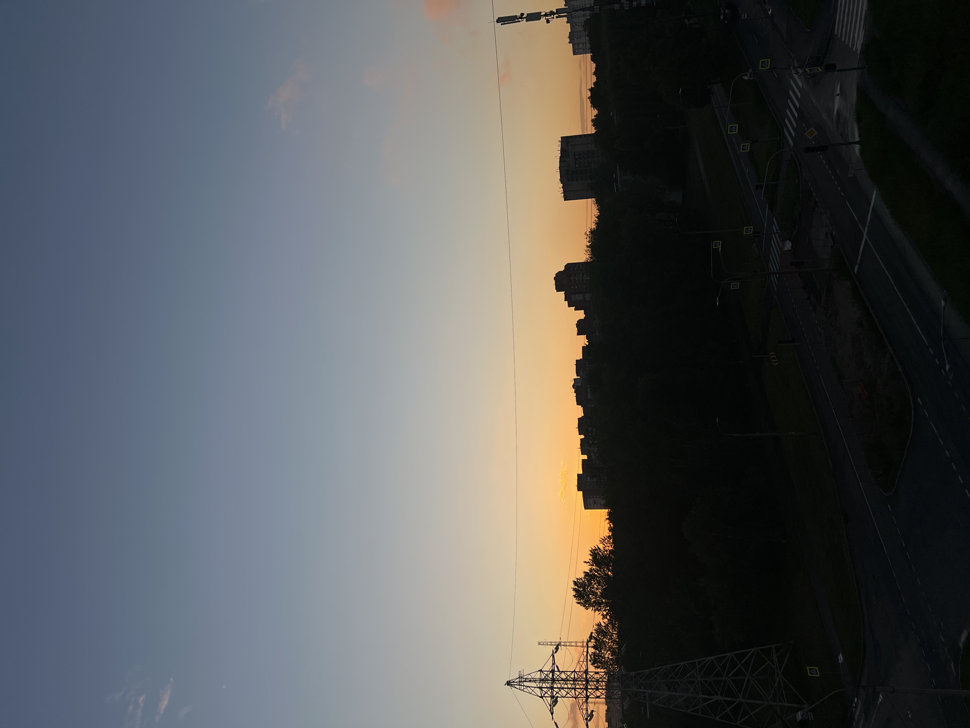 This is a vertical, outdoor, medium-shot, high-angle photograph taken at dusk in Saint Petersburg, Russia. The sky is a gradient of colors, transitioning from a pale blue at the top to a soft orange near the horizon. Silhouetted against the horizon are a row of apartment buildings and a dense line of trees. On the left side of the frame, a large metal power line tower stands tall, with power lines stretching across the image. In the foreground, a dark, paved road intersection is visible, with streetlights and crosswalks. The scene is devoid of human figures.