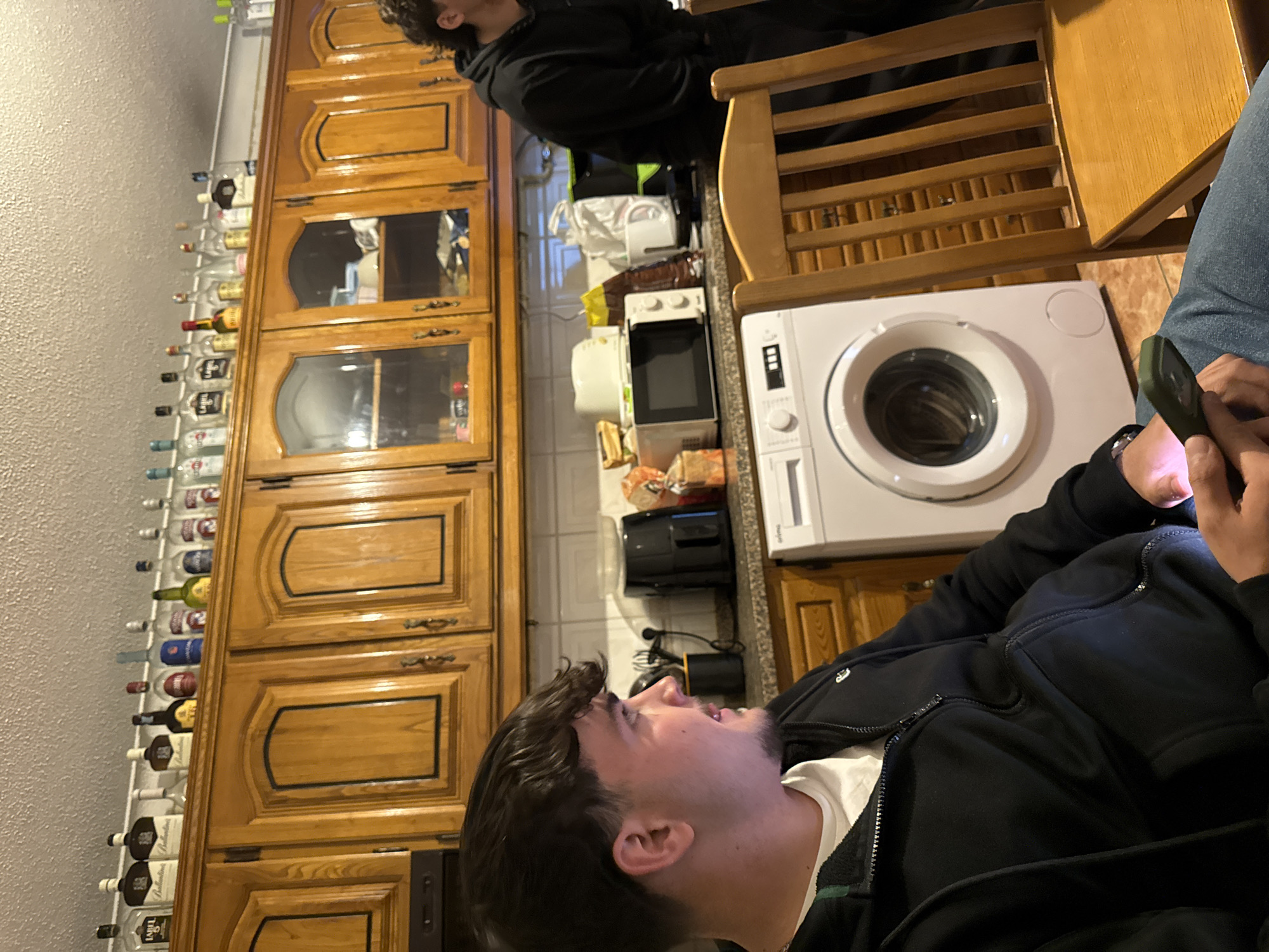 The image shows an interior scene. A person wearing a black jacket and a white t-shirt is holding a phone. In the background, there are wooden cabinets with glass panels displaying bottles, possibly alcohol. A microwave and a white washing machine are visible on the countertop. A wooden chair is also present. It is unknown if the location is in the city of Bragança, Portugal.
