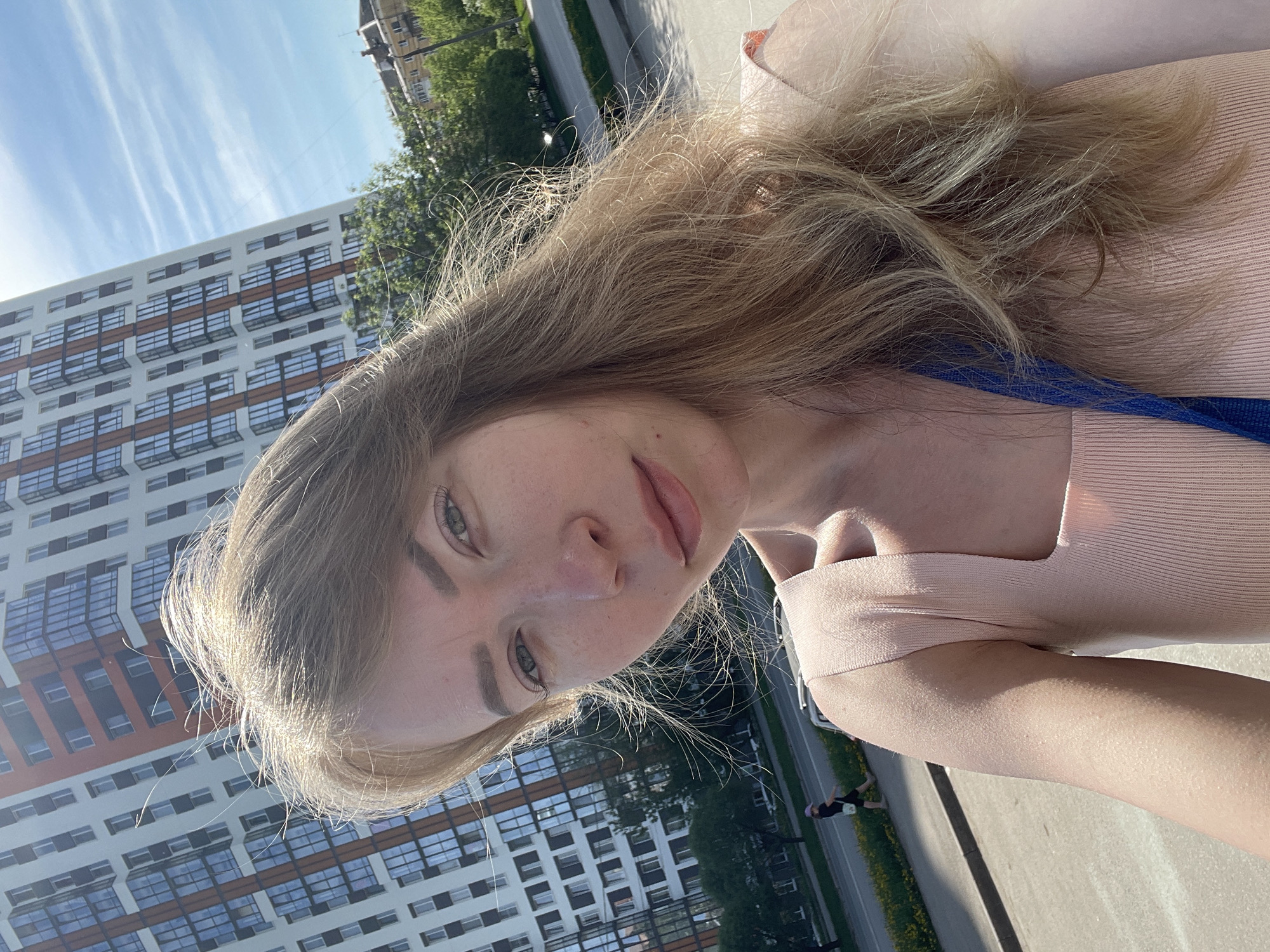 A selfie taken outdoors, featuring a young adult woman with light blonde hair. The camera angle is rotated slightly, showcasing a high-rise building with multiple windows in the background. The woman is wearing a light pink top with a blue strap visible. The background includes trees, a road, and a person walking on the sidewalk. The image was probably taken in Perm, Russia.