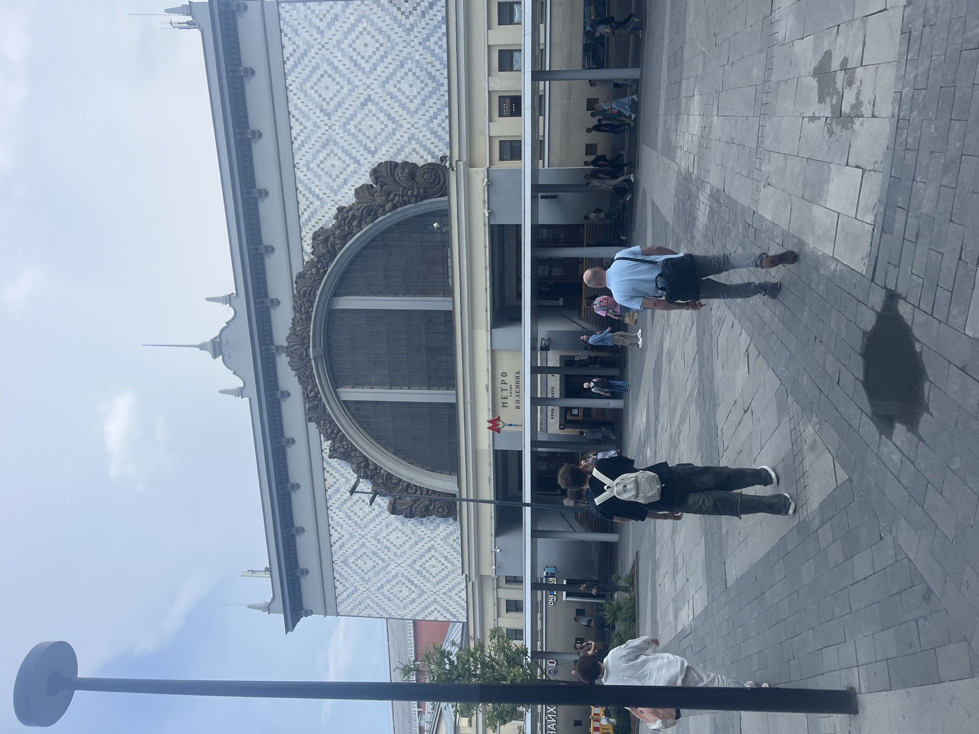 The image shows the facade of the metro station "V.I. Lenin Square" in Moscow, Russia, on a cloudy day. The building features a large arched window with ornate decorative elements and a patterned tile facade. In the foreground, several people are walking on a paved square. One person in the center, seen from behind, wears a black shirt and a white backpack. To their left, another person in a white shirt and light-colored pants also walks away from the camera. To the right, a man in a light blue shirt and grey pants walks towards the station entrance. The ground is paved with grey tiles, with some puddles reflecting the sky. Several trees are visible to the left of the building. The overall atmosphere is that of a public space with moderate pedestrian activity.