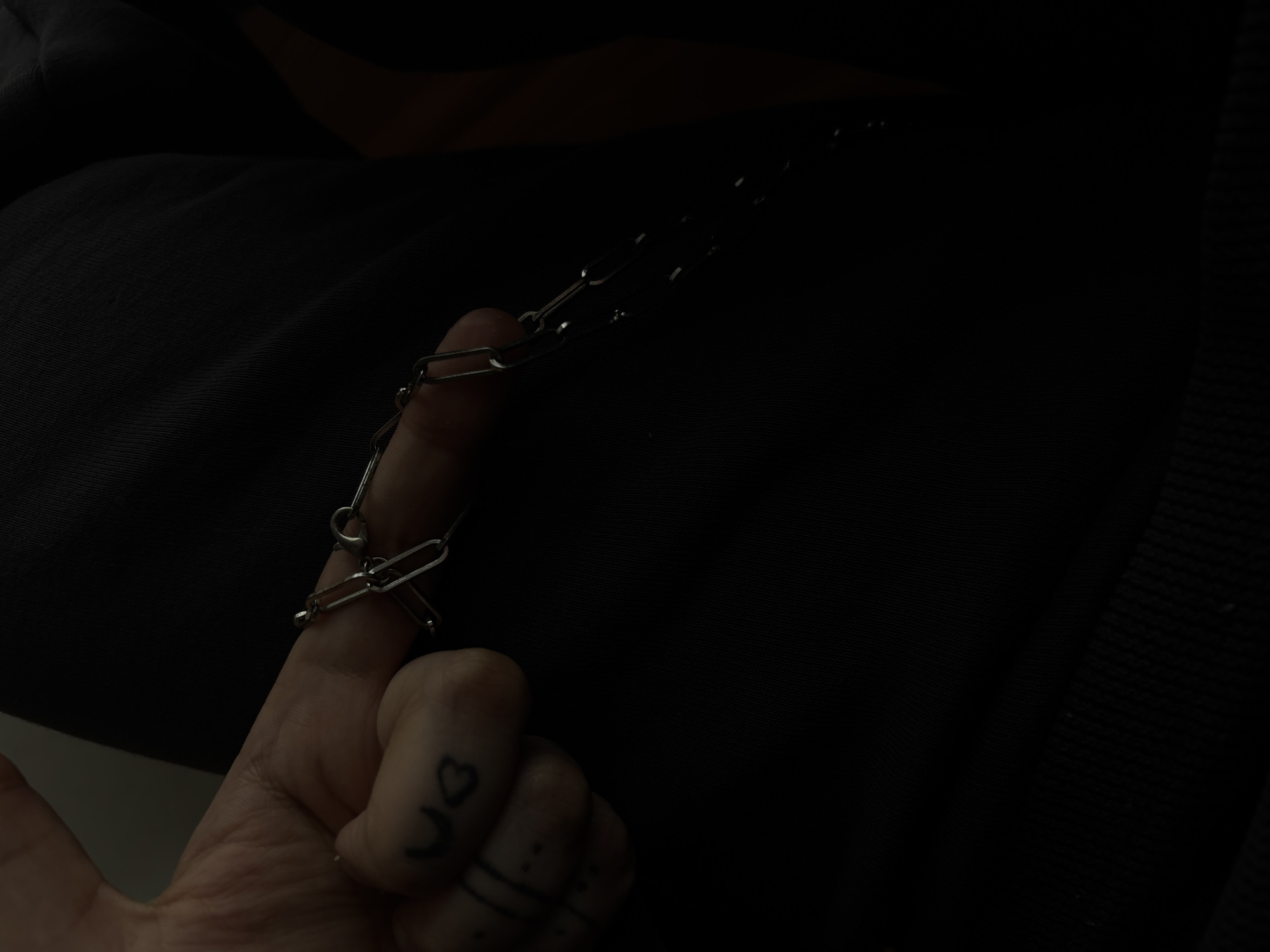 A close-up shot shows a person's hand with tattoos on their fingers, holding a silver chain bracelet with oval links. The bracelet is draped across the index finger. The background is dark and indistinct, appearing to be made of fabric, possibly clothing. The image does not provide any information about the location in the city of Saint Petersburg, Russia.