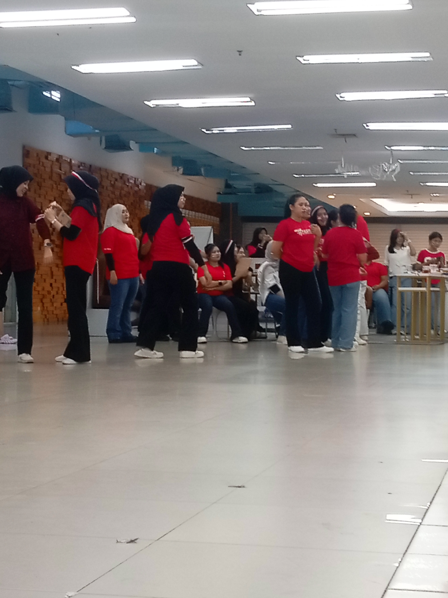 The image shows a group of approximately 15 people, mostly women, gathered in what appears to be an indoor public space, possibly a mall or event hall, in Batam, Indonesia. The lighting suggests fluorescent overhead fixtures. Many of the women are wearing red t-shirts. Several are wearing headscarves, indicating a likely cultural context where this is common. One woman in the foreground, to the left, is wearing a maroon top and black pants. To her right, a woman in a red top and black pants is holding a small object. Further to the right, a woman in a red top and blue jeans is visible. Several other individuals are seen in red tops, some with black pants and some with blue jeans. The background shows a textured wooden wall on the left and a lighter colored wall on the right. There is also a metal structure with shelves in the distance, and a table with items on it. The floor is light-colored and appears to be tiled. The overall scene suggests a casual gathering or event.
