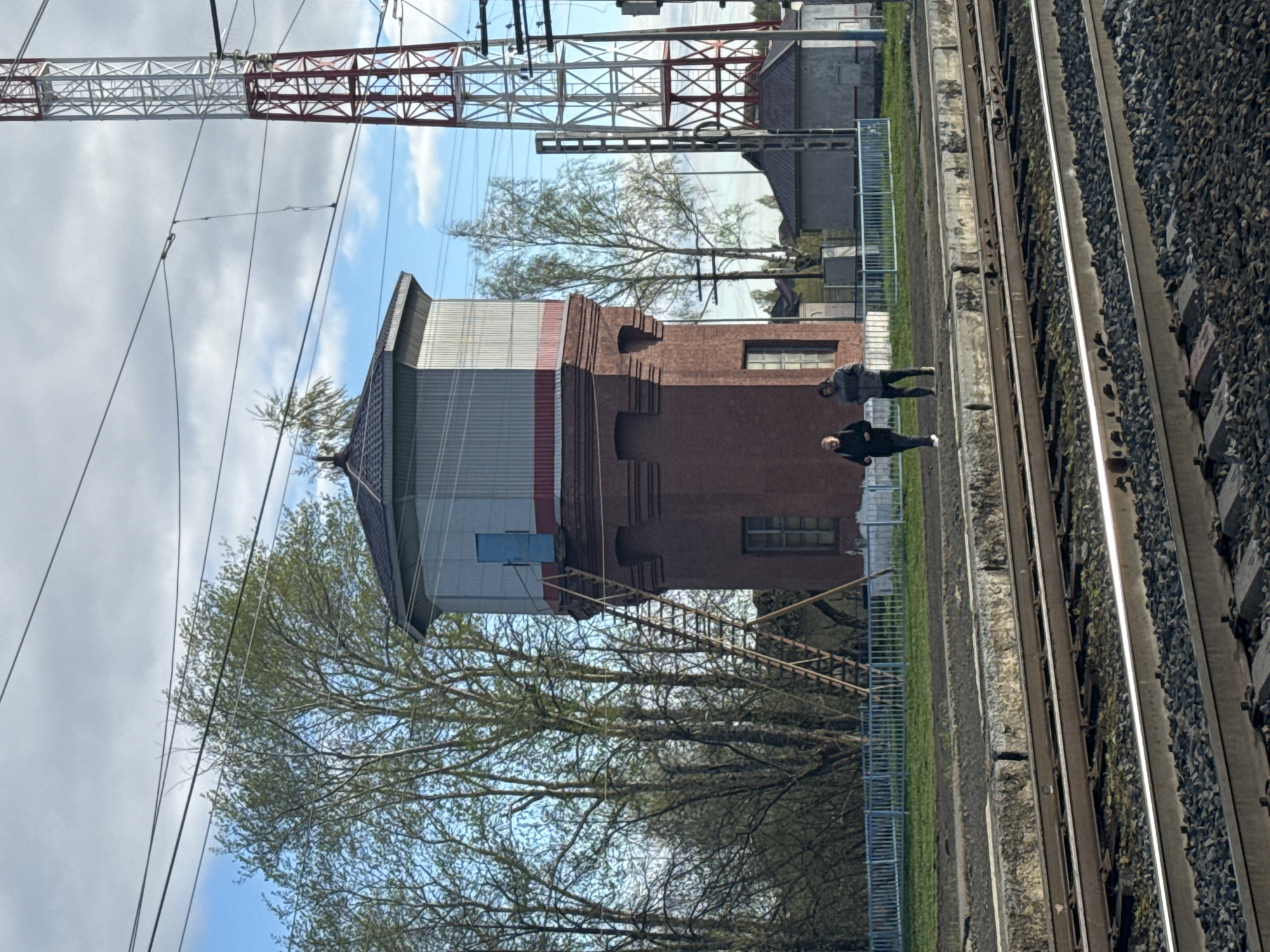 The image captures a scene in Бородулино, Russia, showcasing a water tower constructed from red brick, surmounted by a metallic, multi-tiered structure in gray and white with red bands. The tower is positioned adjacent to a railway line featuring parallel tracks and crushed stone ballast. A green grassy area bordered by a metallic fence separates the tower from the tracks. Two people are walking along a path alongside the railway line. Overhead, a lattice tower carrying power lines is visible against a backdrop of a cloudy blue sky. Trees with budding leaves frame the lower portion of the image, partially obscuring the lower sections of the tower.