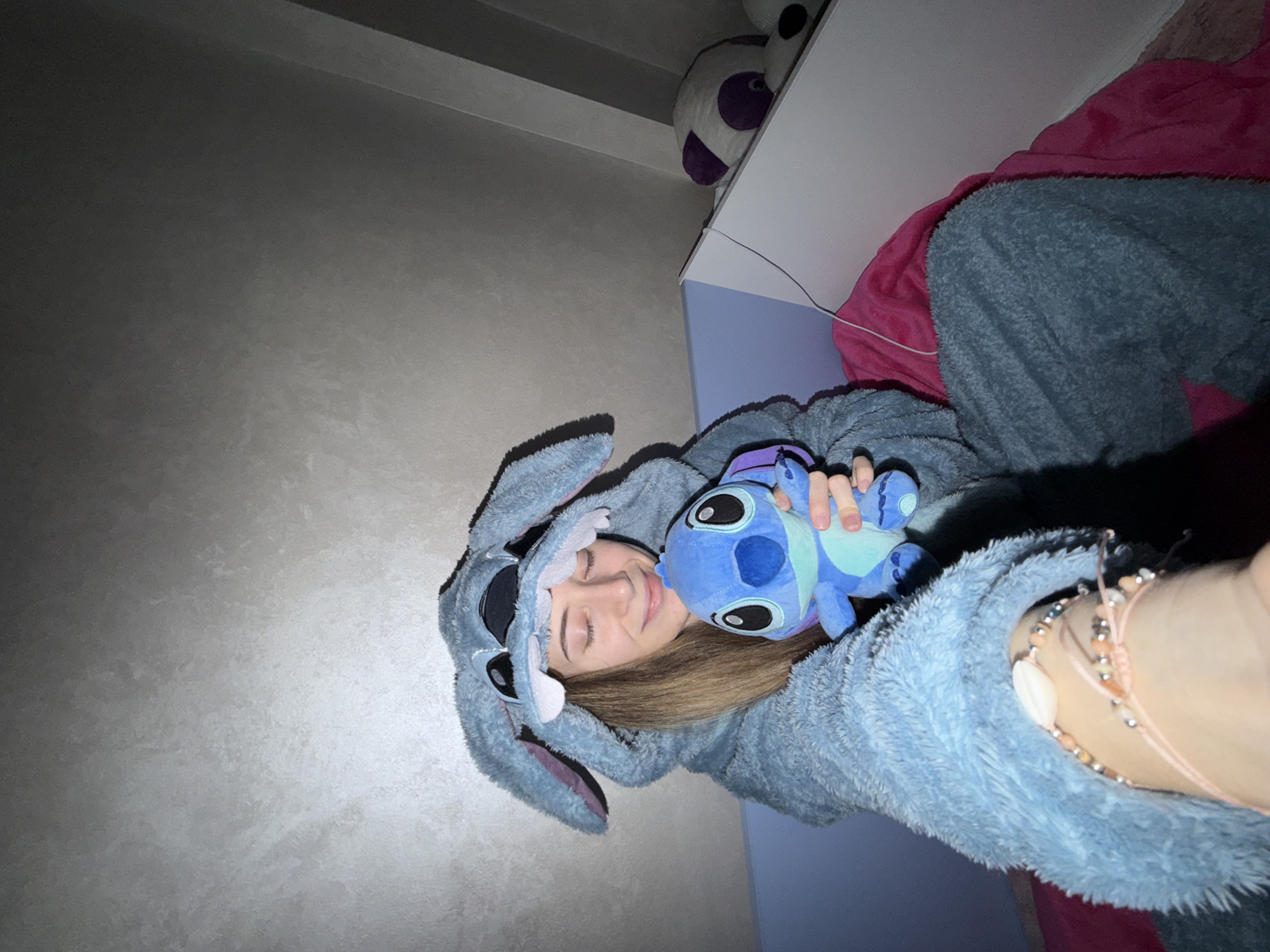 A person is lying on a bed, wearing a light blue, fluffy onesie resembling a cartoon character, and holding a plush toy that resembles the same character. The person is facing upwards towards the camera, smiling with their eyes closed. A portion of their arm and hand are visible in the foreground, with several bracelets on the wrist. The bed has a pink blanket. A nightstand is partially visible with a stuffed panda on top. The room has light gray walls. The location is unknown and is not verifiable as the city of Plovdiv, Bulgaria.