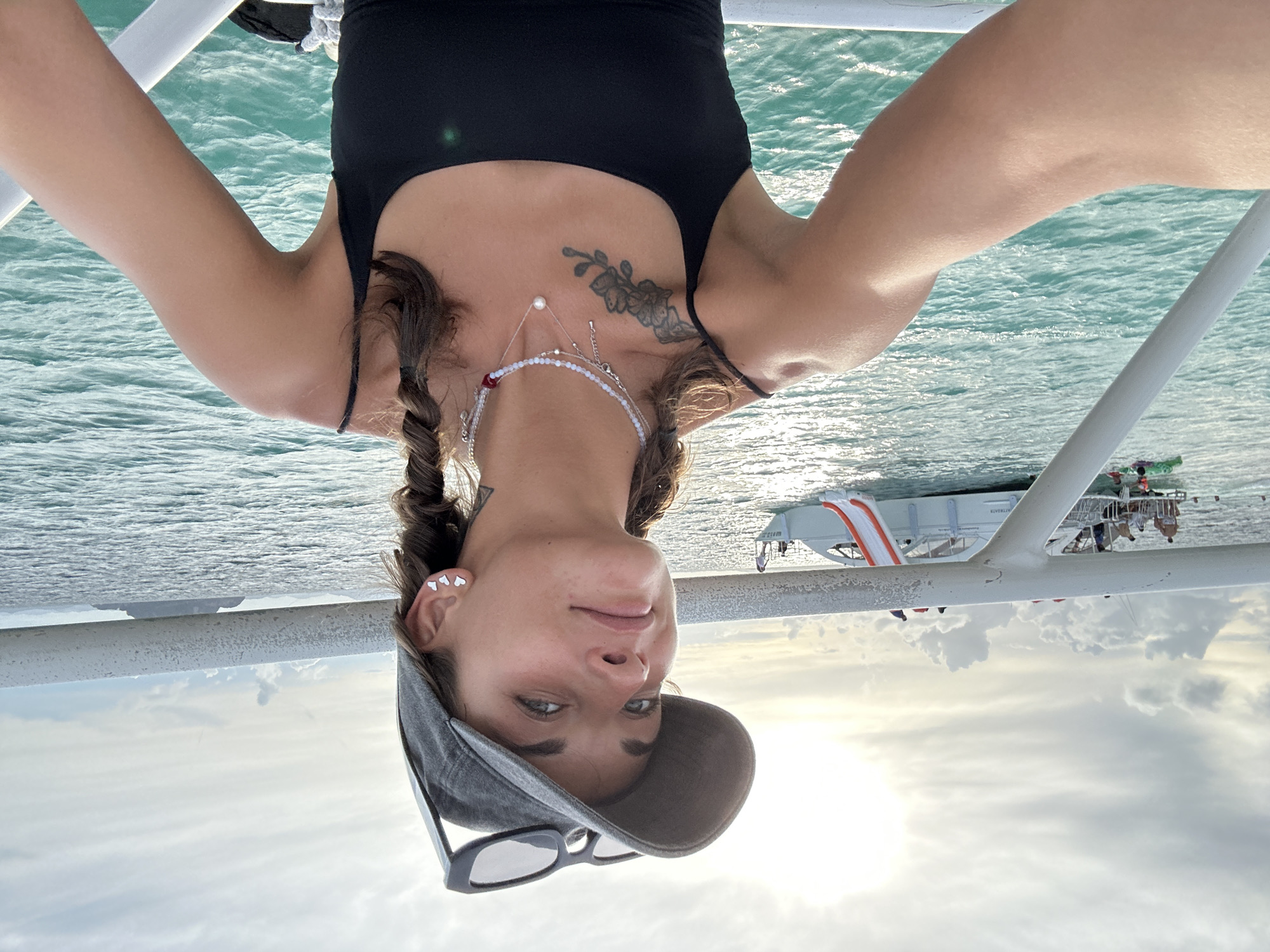 A woman with braided brown hair and a grey baseball cap tilted back poses for a selfie on a boat. She is wearing a black tank top and several necklaces. She has a tattoo of flowers on her left shoulder. In the background, other boats and people are visible in the turquoise water. The sky is overcast with the sun peeking through. The scene appears to be a tourist activity in a tropical location.