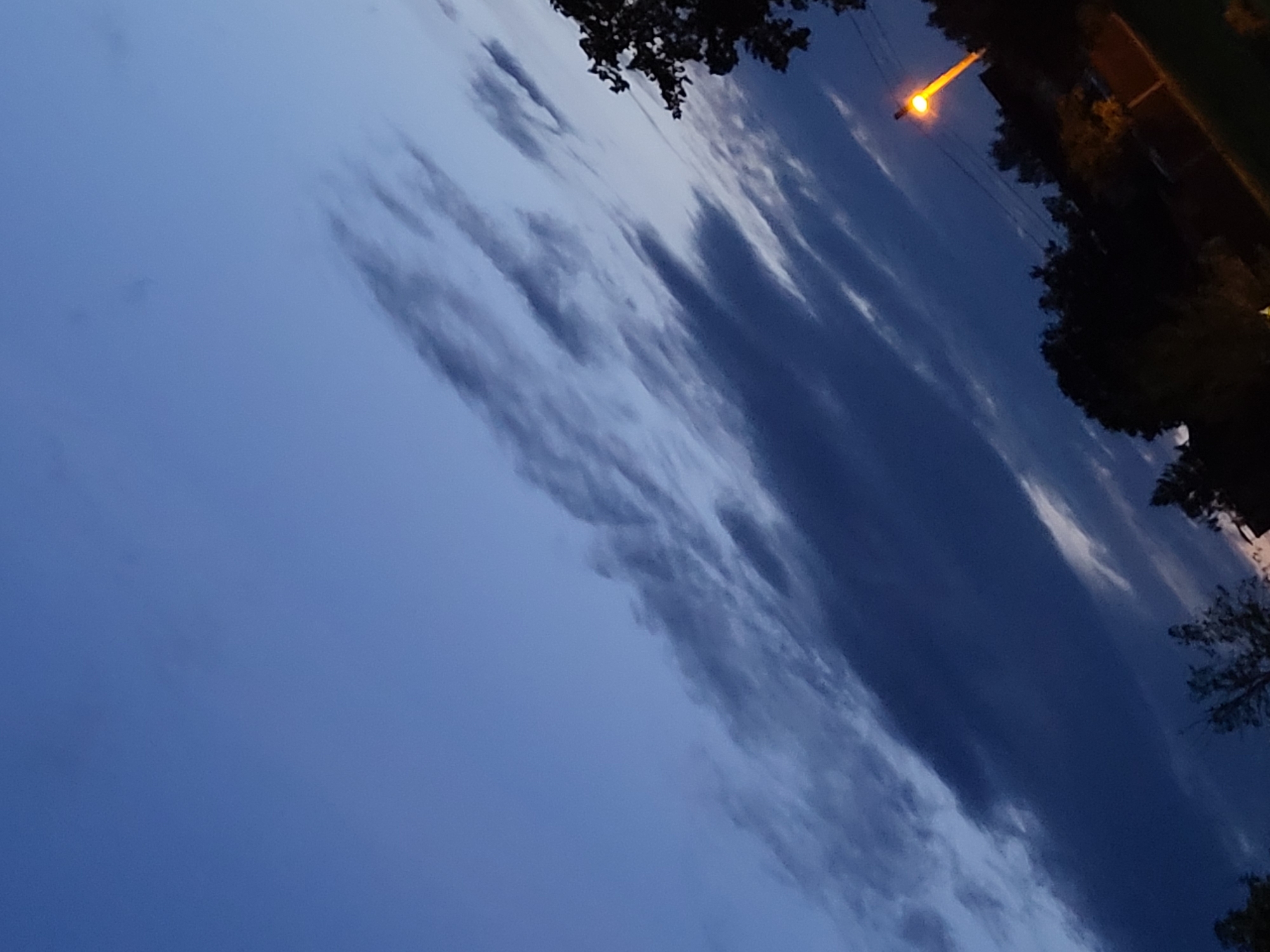 The image depicts a sky with wispy clouds at dusk, angled diagonally from the bottom left to the top right. The sky transitions from a lighter blue at the top to a darker shade towards the bottom. In the lower right portion of the image, a streetlamp with a bright orange light illuminates the scene. Silhouetted against the sky are the tops of trees and what appears to be the roofline of a building. The image suggests a twilight setting in an outdoor environment, possibly a park or residential area within Jelgava, Latvia.