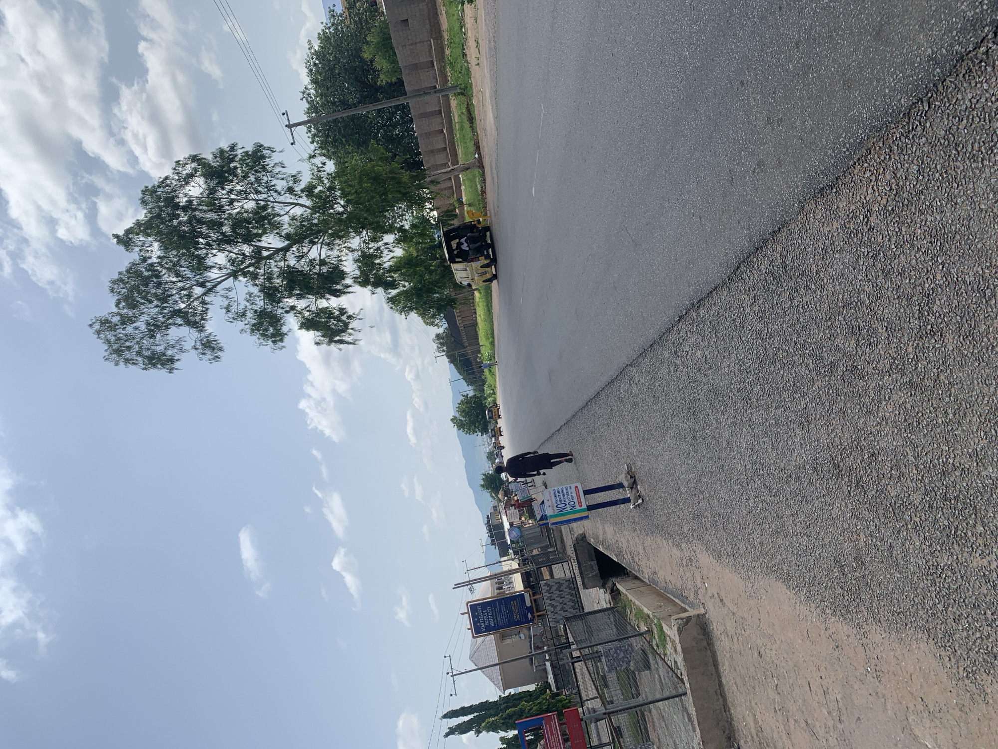 A wide asphalt road stretches into the distance in Jalingo, Nigeria, under a bright, partially cloudy sky. The road has a distinct texture change, with a rougher, gravelly surface on the left and a smoother, dark asphalt on the right. A white drainage ditch runs along the left side of the road, next to a dirt path.
On the left, a metal fence encloses an area with buildings and signage, including a sign for "STAR EXCLUSIVE HOTELS & HOSPITALITY." A person in dark clothing walks away from the camera on the sidewalk. Two blue and white signs are planted in the ground on the sidewalk, one of which reads "NO WAITING NO PARKING."
Further down the road, a yellow van is parked with its rear open. Several people are visible in the distance, as are other vehicles on the road, suggesting ongoing traffic. The landscape beyond the road features green trees and a low brick wall, with distant hills or mountains under the expansive sky. A lone utility pole with wires is visible near a large tree.
