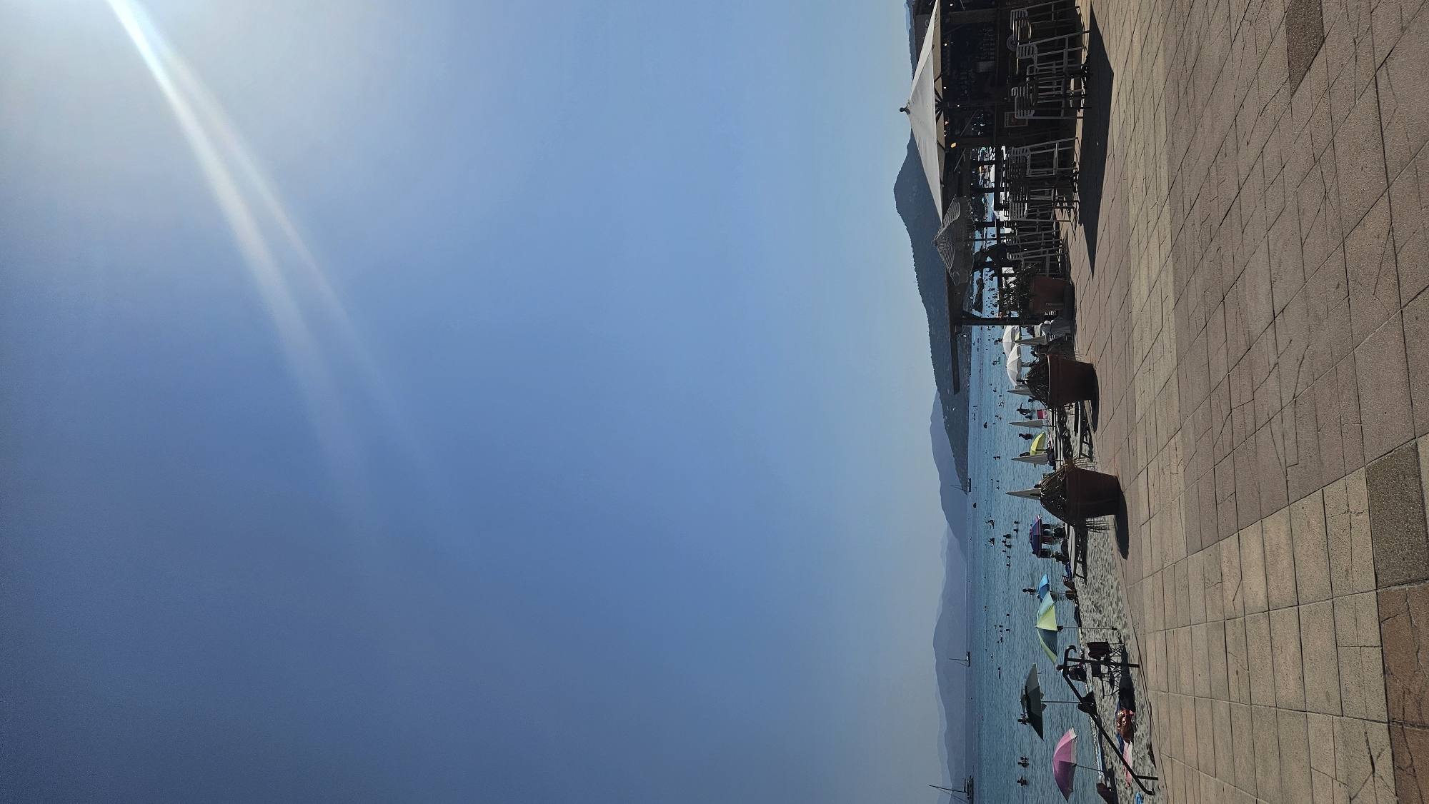 The image captures a bright, sunny day at a beachside location in L'Île-Rousse, France. The scene is divided into three main horizontal sections: the sky, the sea, and the beach/promenade area.
The sky is a clear, deep blue, with a bright sun positioned in the upper right quadrant, casting a lens flare across the sky. Faint, wispy clouds are visible in the distance, subtly catching the sunlight.
The sea is a vibrant turquoise, stretching to the horizon where it meets a range of low, hazy mountains. Several sailboats and other vessels are visible on the water. The water is populated with numerous people swimming and enjoying various water activities.
The foreground features a sandy beach lined with beach umbrellas in various colors, including pink, green, and white. Sun loungers and chairs are scattered on the sand. Adjacent to the beach is a paved promenade constructed of large, rectangular stone tiles. Along the edge of the promenade, a beachside bar or restaurant is set up with numerous high stools and a counter. Large potted plants are placed strategically. The architecture of the bar area, with its thatched and white canvas roofs, suggests a relaxed, tropical ambiance. The overall scene depicts a busy and lively summer day at a popular coastal resort.