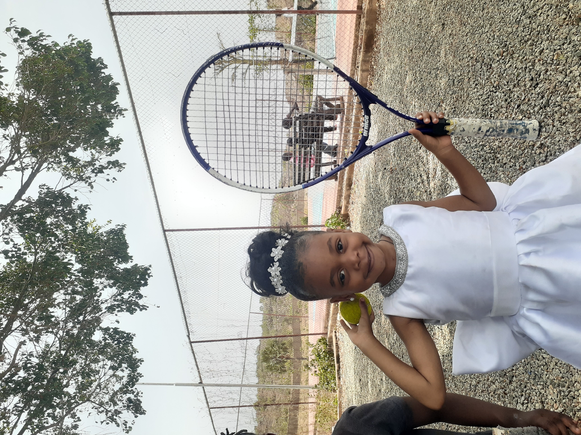 A young girl stands on a gravelly surface near a chain-link fence. She wears a white dress and holds a tennis ball and a tennis racquet. Beyond the fence, there are trees and a concrete area. The location is in Jos, Nigeria.