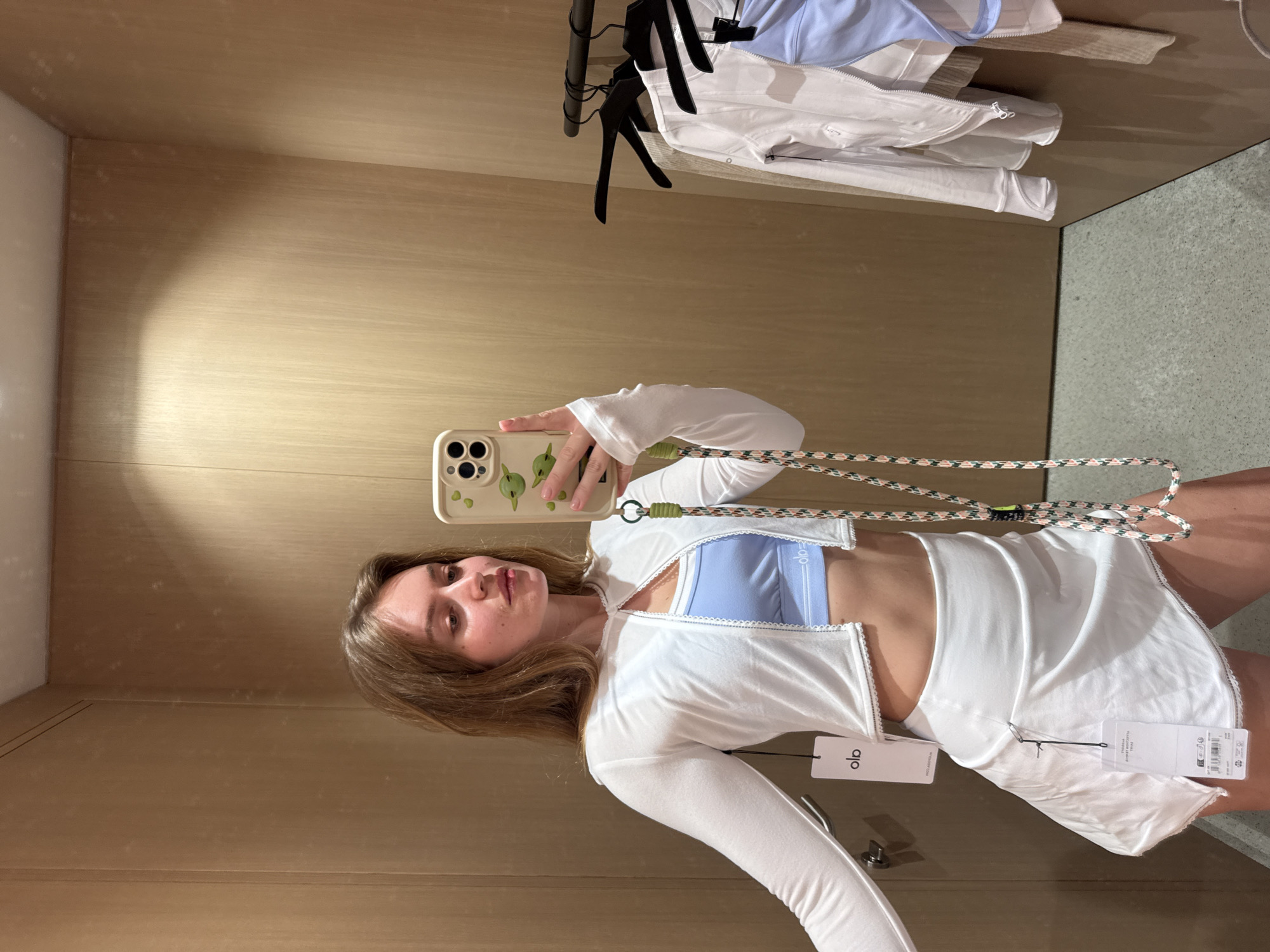 A young woman is captured in a mirror selfie within what appears to be a fitting room, likely in a retail store in Rome, Italy. She has long, light brown hair and a neutral expression, looking directly at the camera. She is wearing a light blue sports bra, a white, long-sleeved cropped jacket or shrug, and a white activewear skirt or shorts. The jacket and skirt both feature an "alo" brand tag, visible near her waist. The "alo" logo is also seen on the blue sports bra.
She holds a light-colored smartphone with a light green case featuring a distinct pattern, possibly of avocado halves. A patterned strap hangs from her phone. In the background, behind her, a clothes rack holds several white and light blue garments on black hangers. The fitting room features light wood-paneled walls and a light gray, textured floor. The image is taken from a slightly upward angle, capturing her mid-body and the details of her outfit and the immediate surroundings.