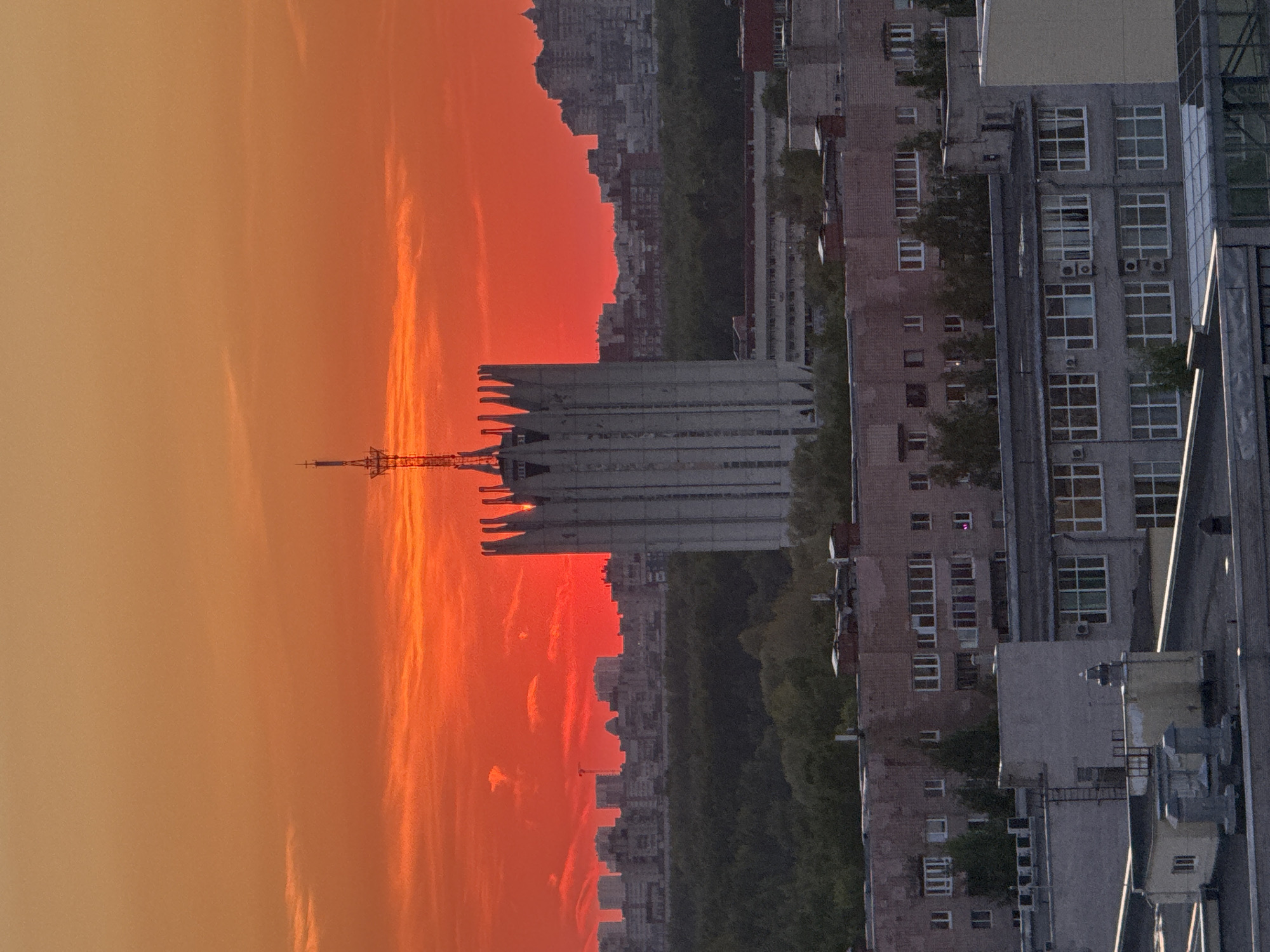 The image shows a sunset view in Saint Petersburg, Russia. The sky is an orange-red gradient, with a large, modern building featuring a spire silhouetted against the horizon. The building appears to be located amidst a cityscape, with other buildings of varying heights and styles visible in the background. Some trees are visible between buildings. The photo is taken from a high vantage point, likely from within another building. A neighboring building is partially visible on the right side of the frame, with multiple windows and some air conditioning units. The image is rotated by 90 degrees.