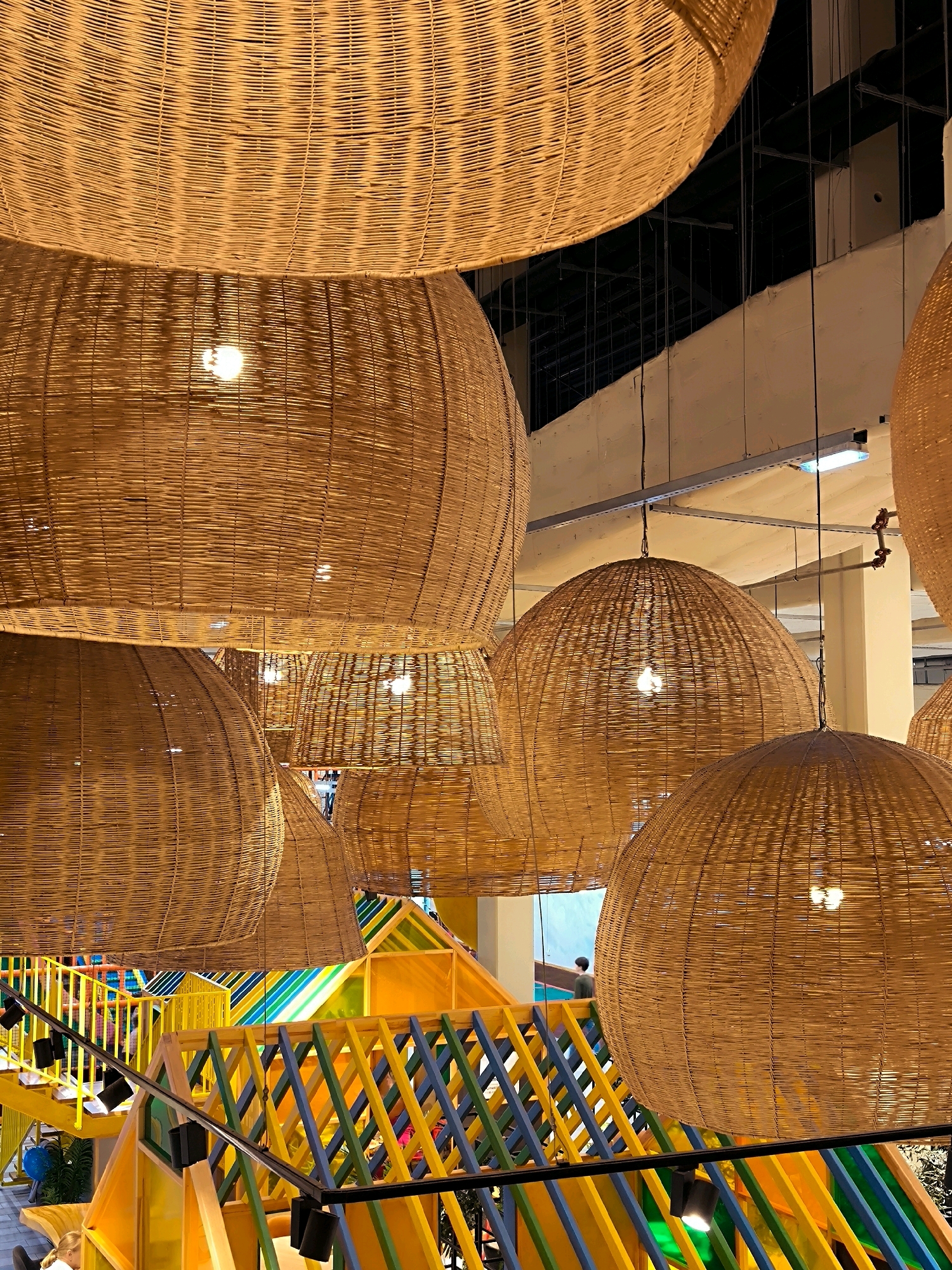 The image displays a collection of large, woven rattan pendant lamps hanging from the ceiling. The lamps are circular and dome-shaped, with intricate weaving patterns that allow light to filter through. Below the lamps, a brightly colored, multi-level children's play structure is visible, featuring yellow, blue, green, and orange elements, including slides and stairs. There are a few people present, including a child at the bottom left and a person walking on the play structure in the background. The environment appears to be indoors, possibly a public space like a cafe, restaurant, or play center, with exposed ceiling beams and lighting fixtures in the background. The overall impression is one of a warm and inviting, yet modern, interior design.
Based on the available information, the location is not specified as Riga, Latvia. Therefore, no information regarding this city can be provided.