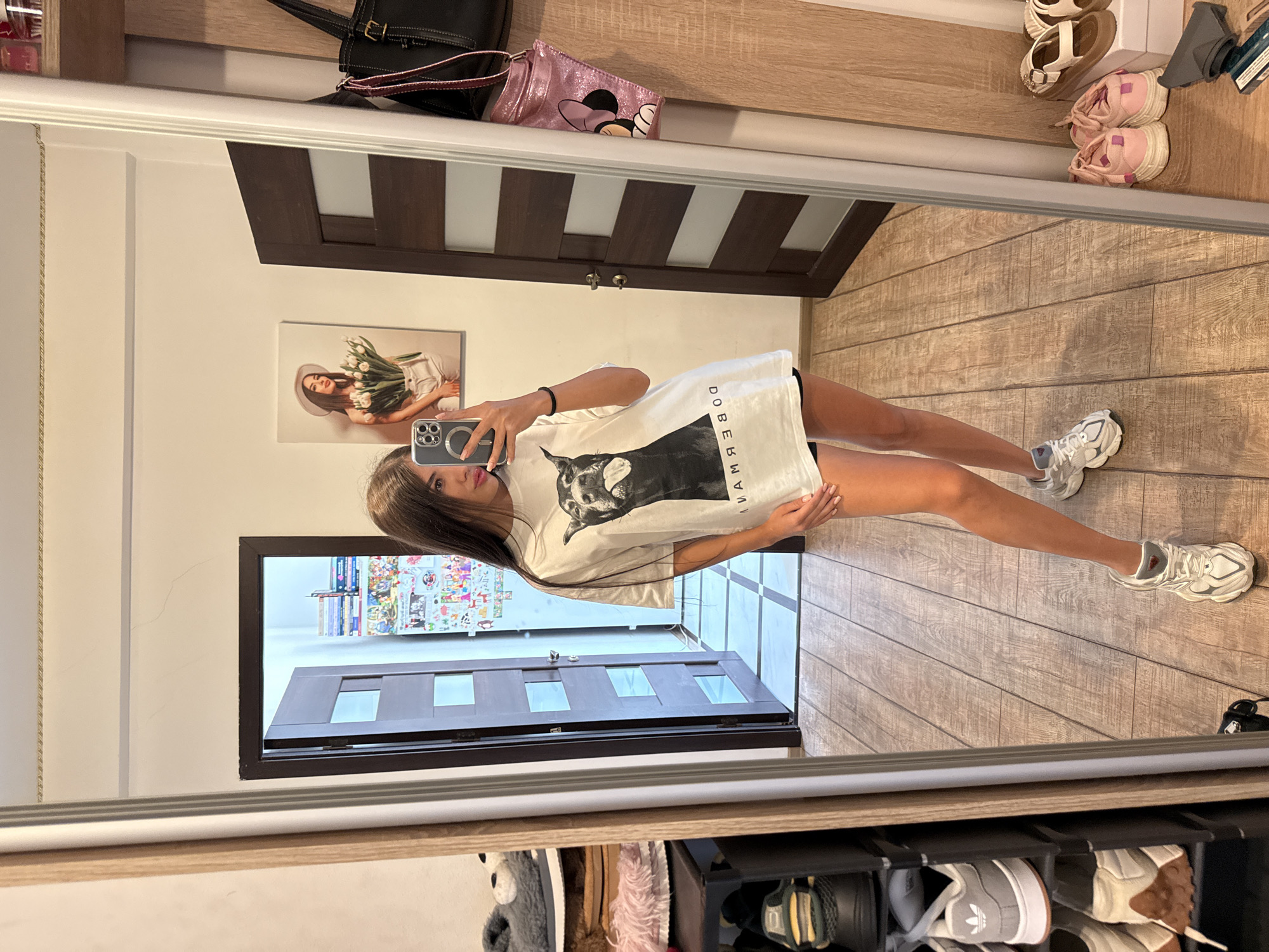 A young woman with long dark hair takes a mirror selfie in a room with wooden floors and doors. She is wearing an oversized white t-shirt with a large black and white graphic of a dog on the front, and black shorts. She has on white and gray athletic shoes. In the reflection, a shoe rack with various shoes is visible to her left. To her right, a dark brown door and a beige door frame are visible. A framed picture of a woman holding a bouquet of tulips hangs on the wall above the beige door frame. A black handbag and a pink sparkly purse with a cartoon character are hanging on the right side of the room. The room appears to be a bedroom or dressing area.