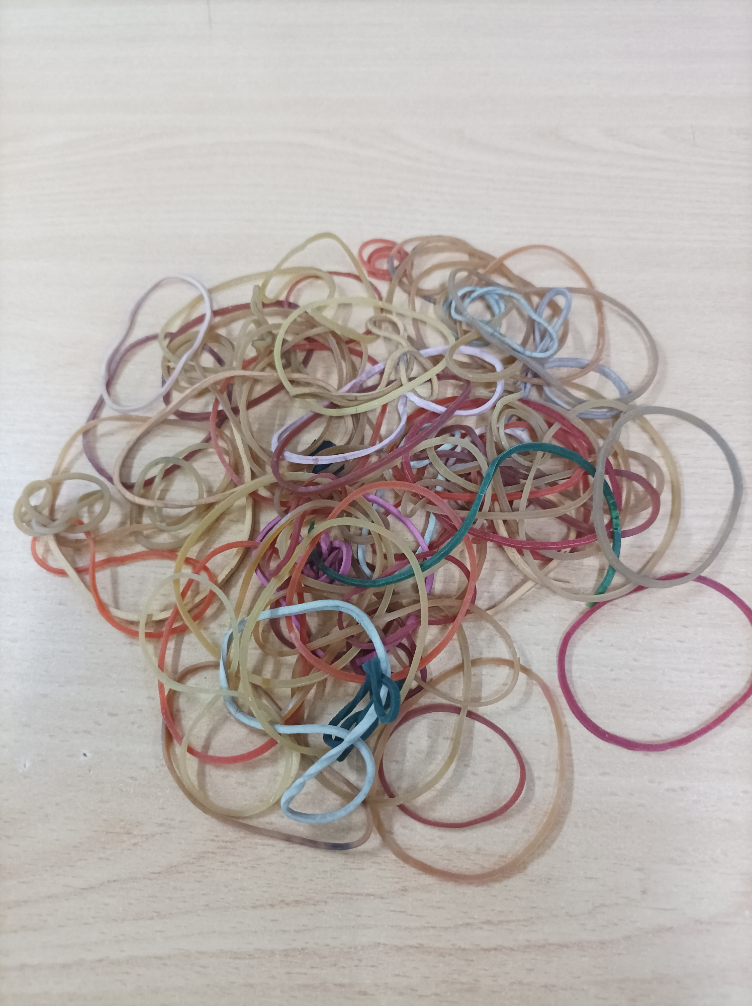 A pile of colorful rubber bands is scattered on a light-colored wooden surface. The rubber bands are a mix of natural tan, red, blue, green, and purple. The surface appears to be a tabletop or desk. There are no people or animals visible in the image. The lighting is natural, casting soft shadows. The image is taken from a slightly elevated angle looking down at the rubber bands. The scene does not depict any specific location within the city of Nganjuk, Indonesia.