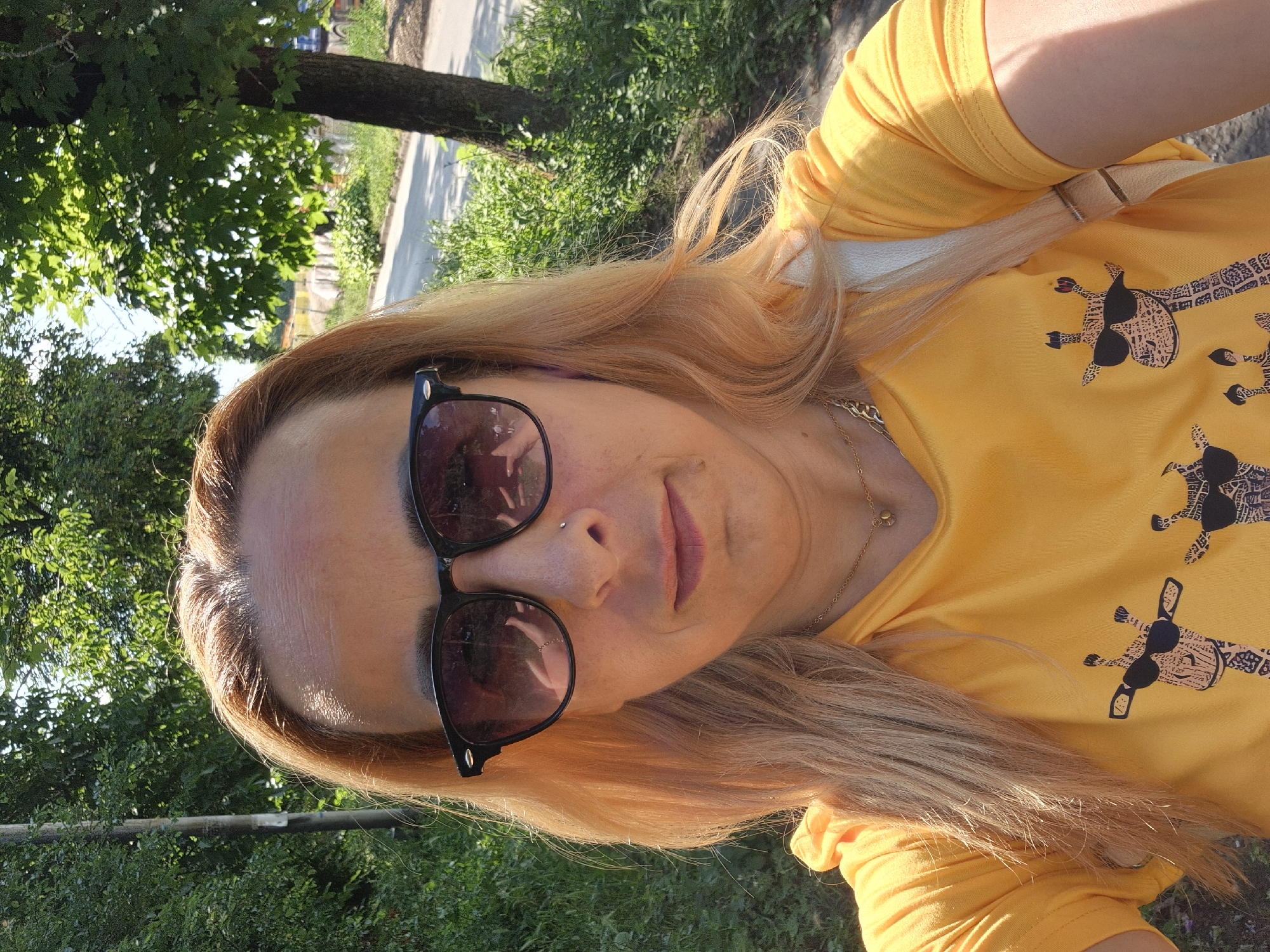 A selfie-style portrait captures a woman outdoors, tilted approximately 90 degrees clockwise. The woman has long, light brown to blonde hair, which flows down and over her shoulder, illuminated by sunlight. She is wearing dark-rimmed sunglasses that obscure her eyes, and a small stud nose piercing is visible on her right nostril. Her lips are lightly colored, showing a subtle smile.
She is dressed in a bright yellow short-sleeved t-shirt, which features a repeating black graphic design of giraffes. Each giraffe in the pattern is depicted wearing sunglasses and a bow tie or scarf with intricate patterns. A thin, gold-colored chain necklace is visible around her neck.
The background consists of green trees, lush foliage, and what appears to be a paved road or path under dappled sunlight. The bright lighting suggests it is daytime, with clear shadows indicating sunny weather. The overall impression is a casual outdoor snapshot. No discernible text is visible in the image.