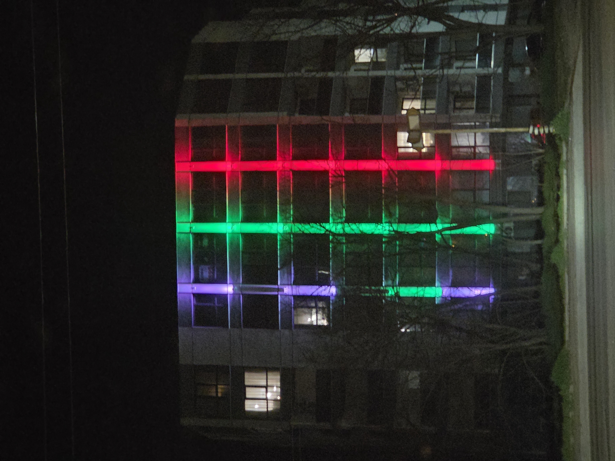 A building illuminated with red, green, and purple lights at night. The location might be in Varna, Bulgaria based on the prompt, but is impossible to determine this with certainty. The lights shine horizontally across the building. Bare tree branches are visible in front of the building. Utility lines are seen on the left side of the image.